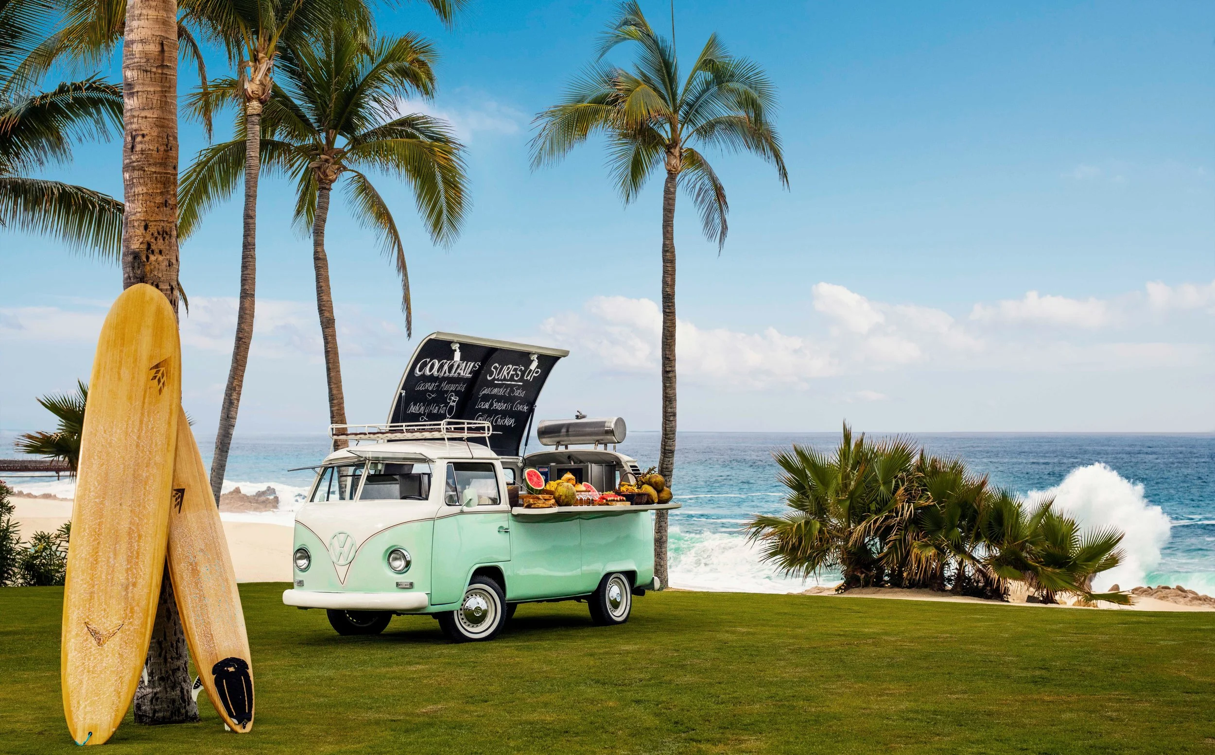 Beach scene with palm trees, a vintage Volkswagen van converted into a food truck with a chalkboard menu, and surfboards leaning against a tree, overlooking the ocean.