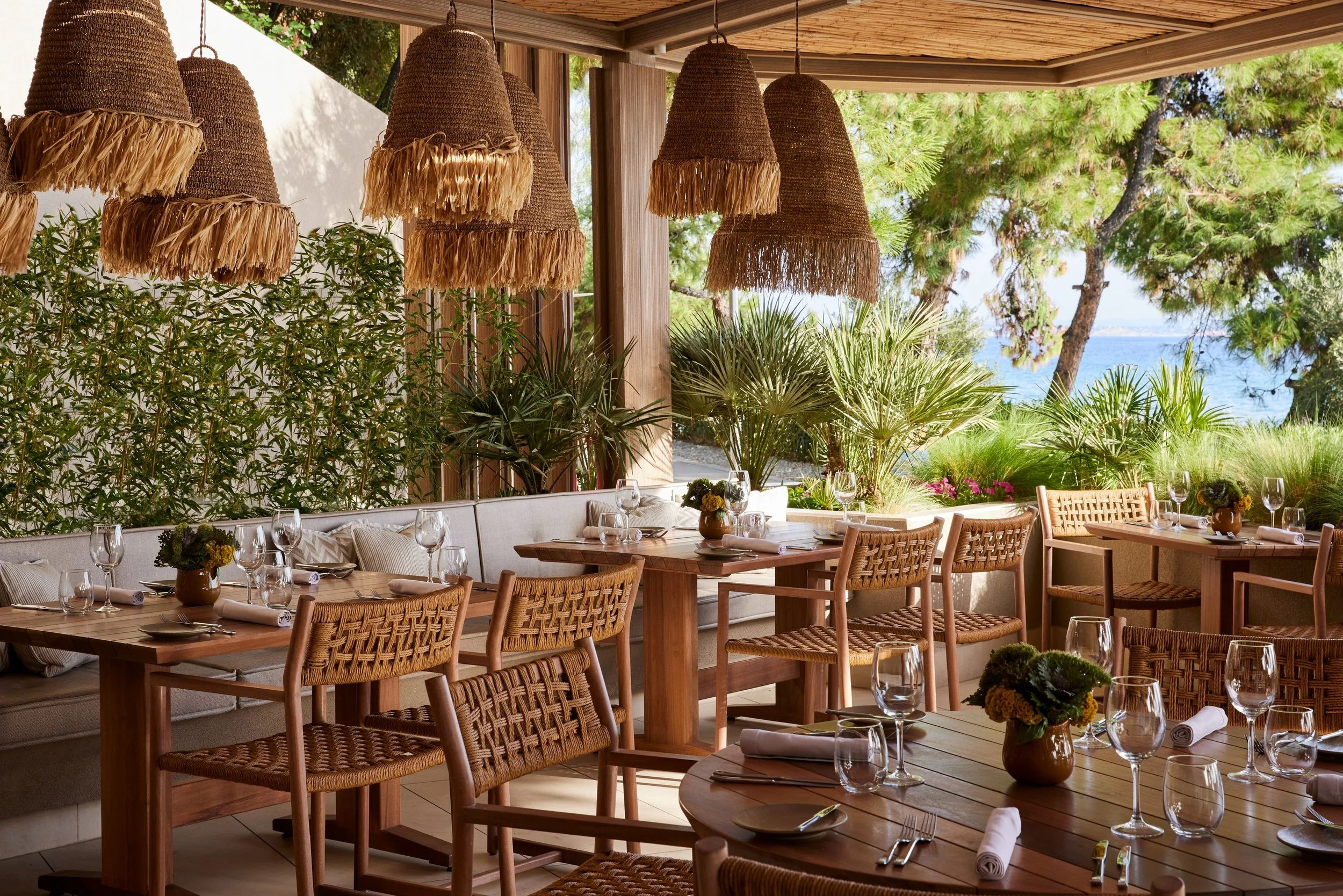 A restaurant with wooden tables and woven chairs, decorated with small flower arrangements. There are hanging woven lamps outside, with trees and greenery in the background and ocean views.