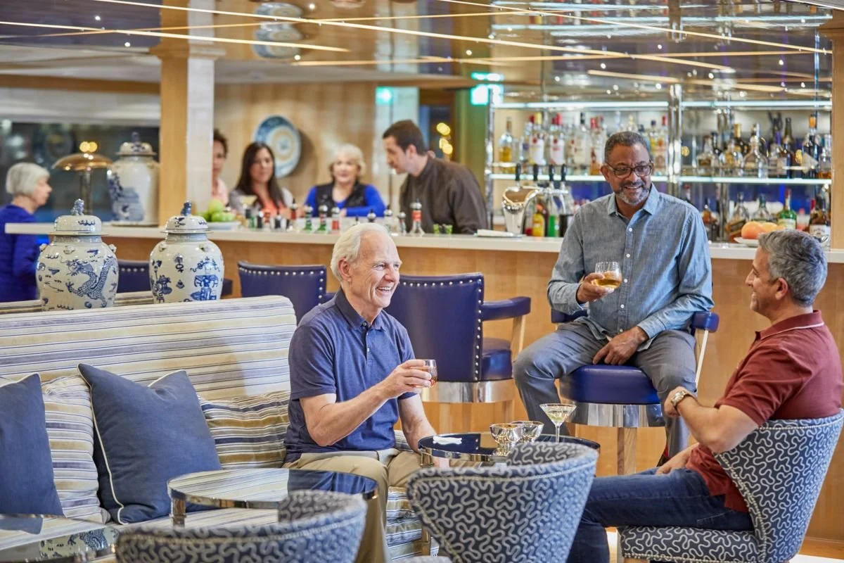 Three men are socializing with drinks in a cozy bar or lounge area, with a bartender and others in the background.