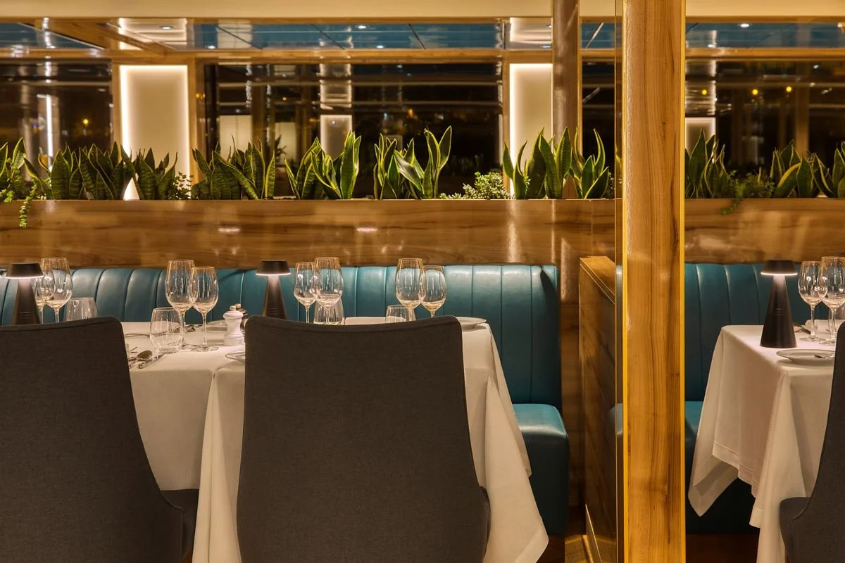 Elegant restaurant interior with white-draped tables set with wine glasses, napkins, and cutlery; teal upholstered banquettes and black chairs, with a decorative wooden partition and lush green plants in the background, illuminated with warm lighting.