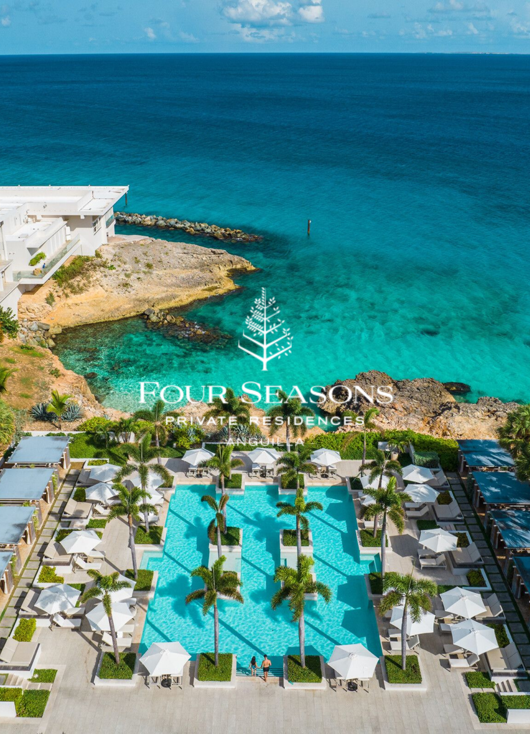 Aerial view of a luxury resort with a large swimming pool surrounded by palm trees, umbrellas, and lounge chairs. In the background, there is an ocean and a rocky shoreline.