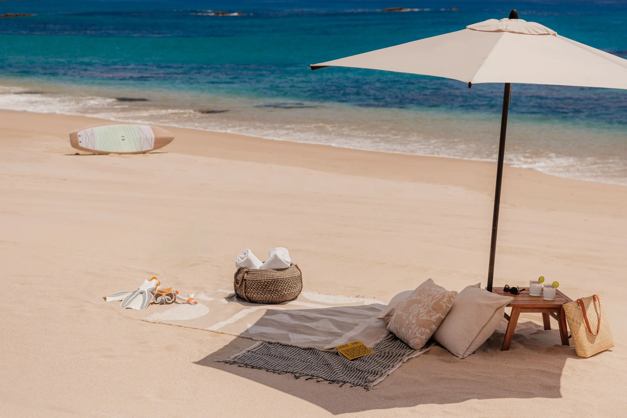 A beach scene with an umbrella, a surfboard, and various beach items on the sand, with the ocean in the background.