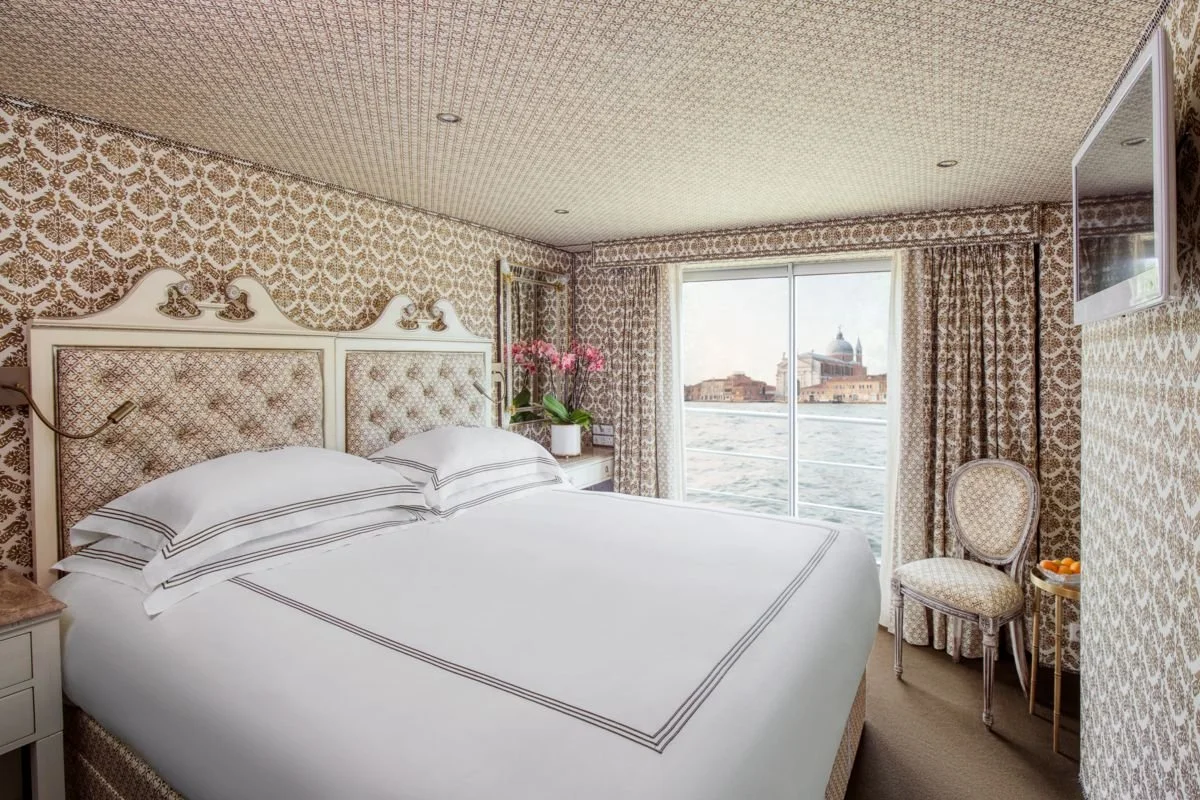 Hotel bedroom with patterned wallpaper and curtains, white bed with striped pillows, a chair, a wall-mounted TV, and a view of a canal or river through large windows.