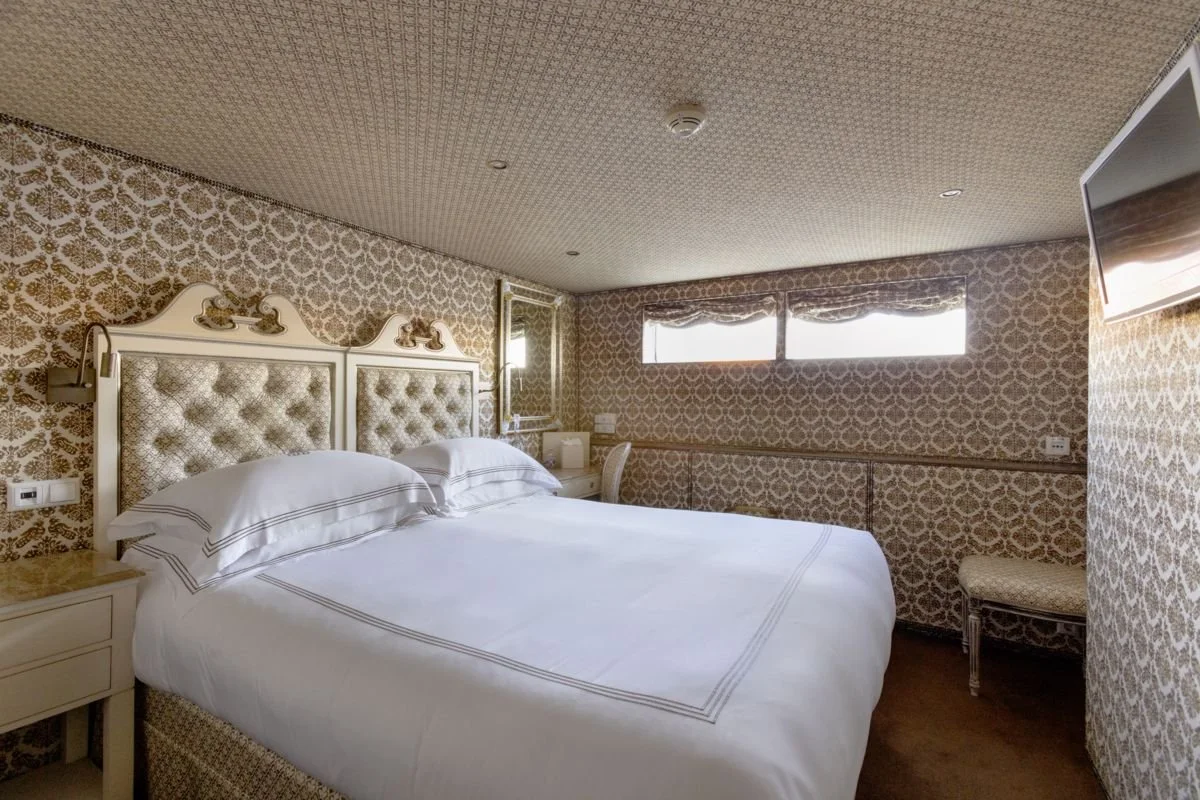 A vintage-style hotel room with patterned wallpaper, a large bed with a tufted headboard, white linens, a nightstand, a wall mirror, and a window with a valance, on the right side a wall-mounted TV, and a small bench.