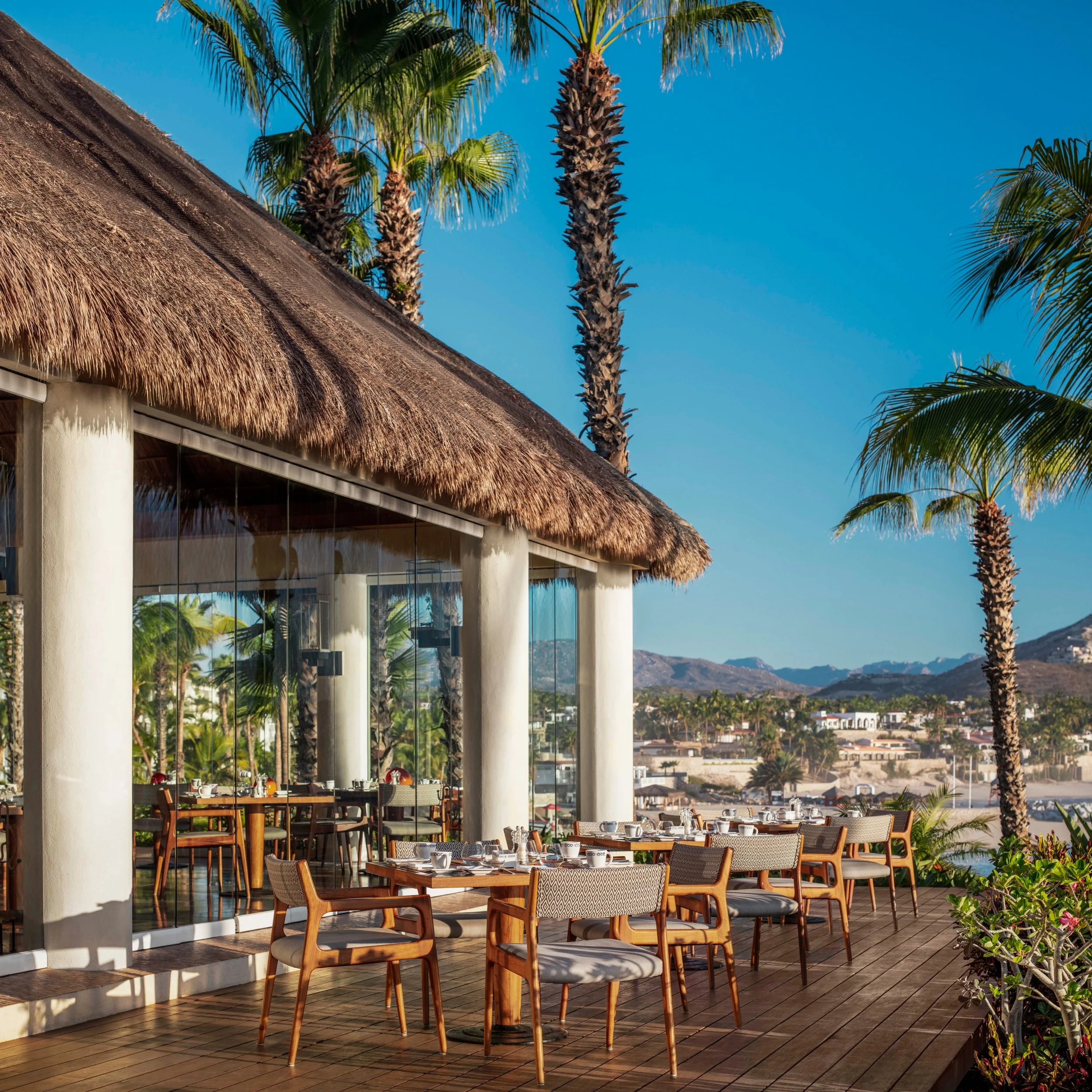 An outdoor patio area with wooden tables and chairs, a thatched roof structure, palm trees, and a view of a cityscape with hills in the distance.