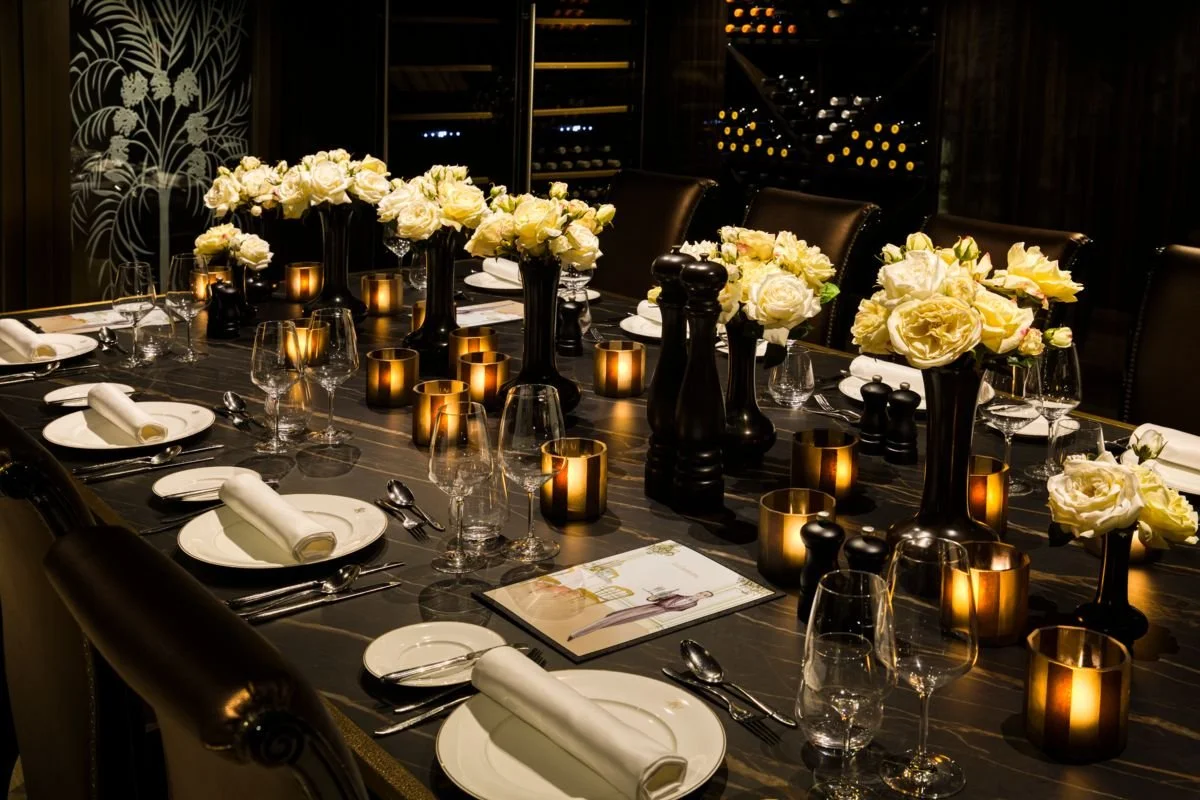 Elegant dinner table setup with white flower centerpieces, lit candles, wine glasses, and place settings with white napkins in a dimly lit wine cellar or private dining room.