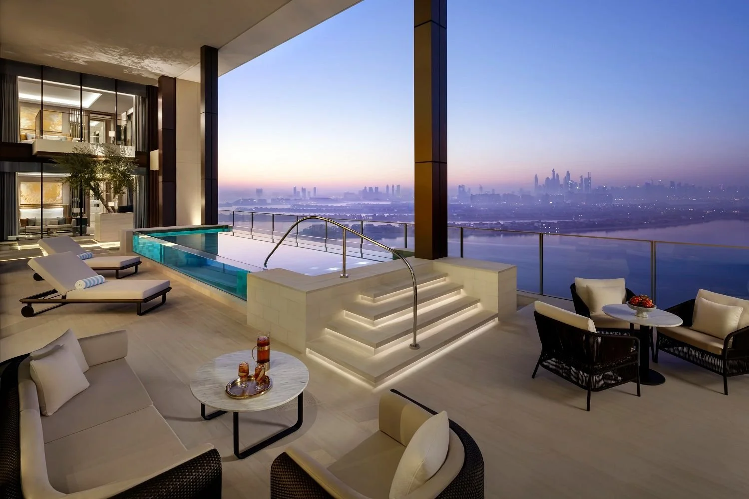 Luxury rooftop balcony with infinity pool, lounge chairs, and outdoor seating area overlooking a city skyline at sunset.