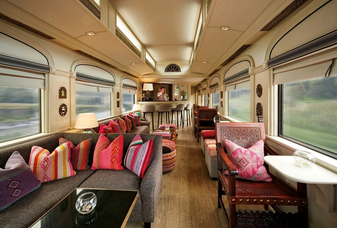 Interior of a vintage train car with large windows, plush seating, colorful pillows, and a bar area in the background with a person standing.