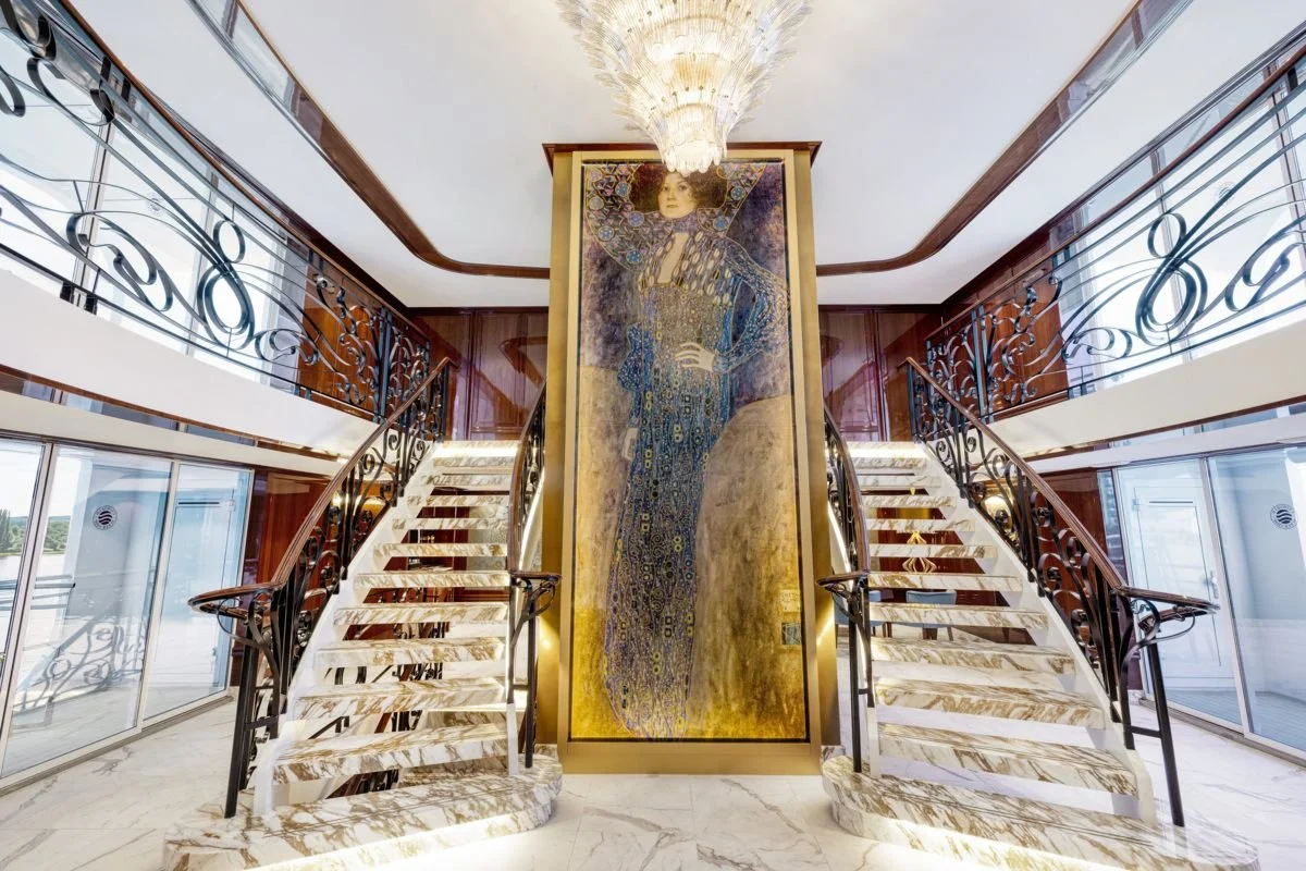 Luxurious interior with two marble staircases leading to a second floor, framed by decorative black wrought iron railings, a large colorful portrait of a woman in a flowing dress in the center, and a grand chandelier hanging from the ceiling.
