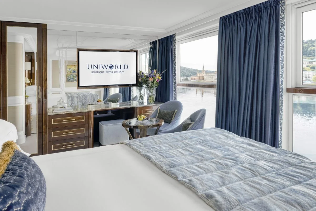 A hotel room with a bed, blue curtains, and large windows overlooking a river and cityscape. There's a TV on a wooden dresser with the logo of Uniworld Boutique River Cruises displayed on the screen. A small round table with a flower arrangement and two chairs are visible near the windows.