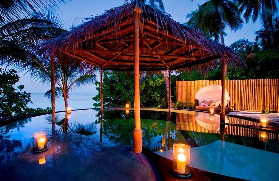 Tropical outdoor lounge area with a thatched-roof gazebo, surrounded by palm trees and lit by candles at dusk.