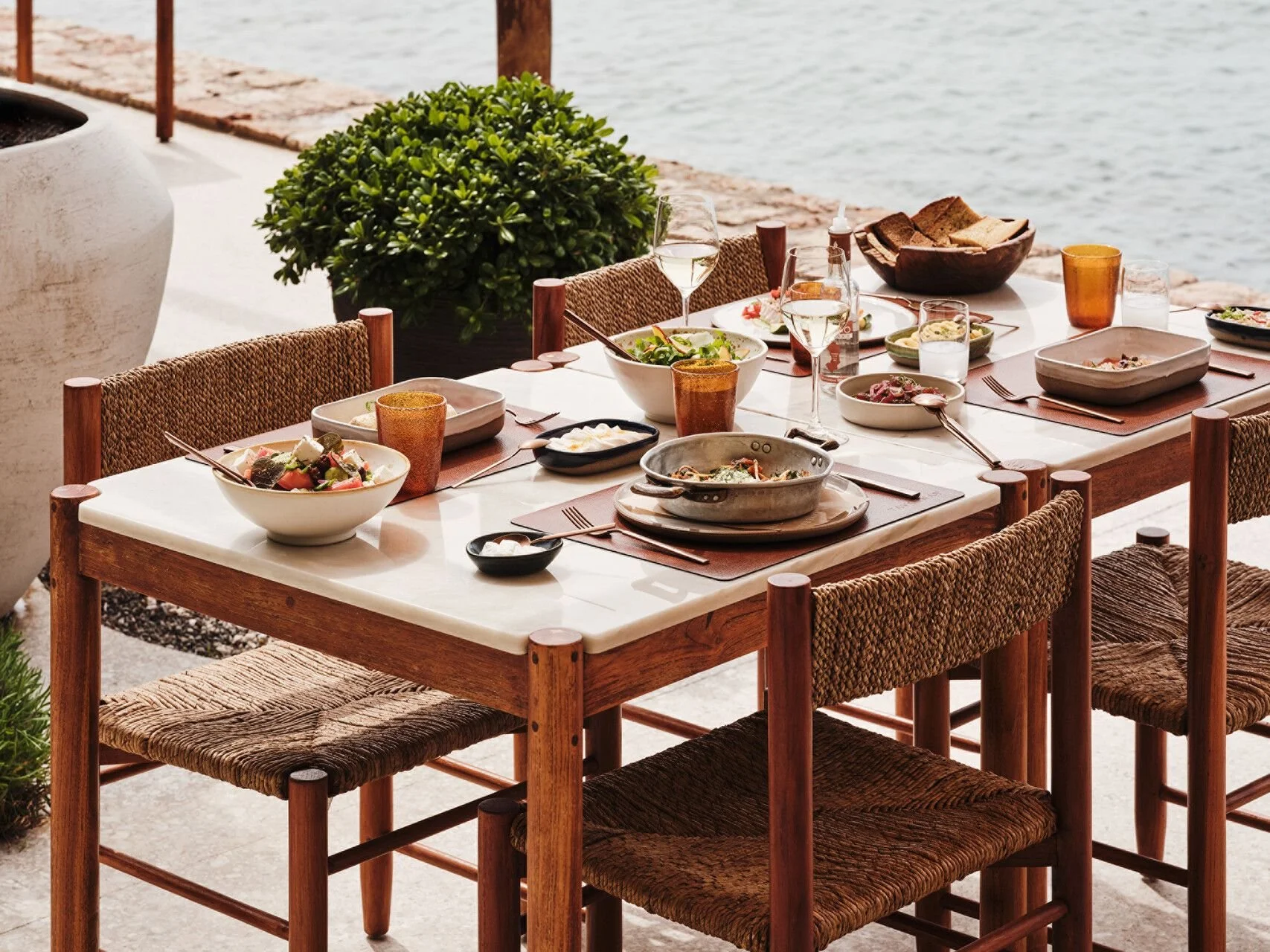 A dining table set outdoors near water, with plates of food, glasses, and a bowl of bread.