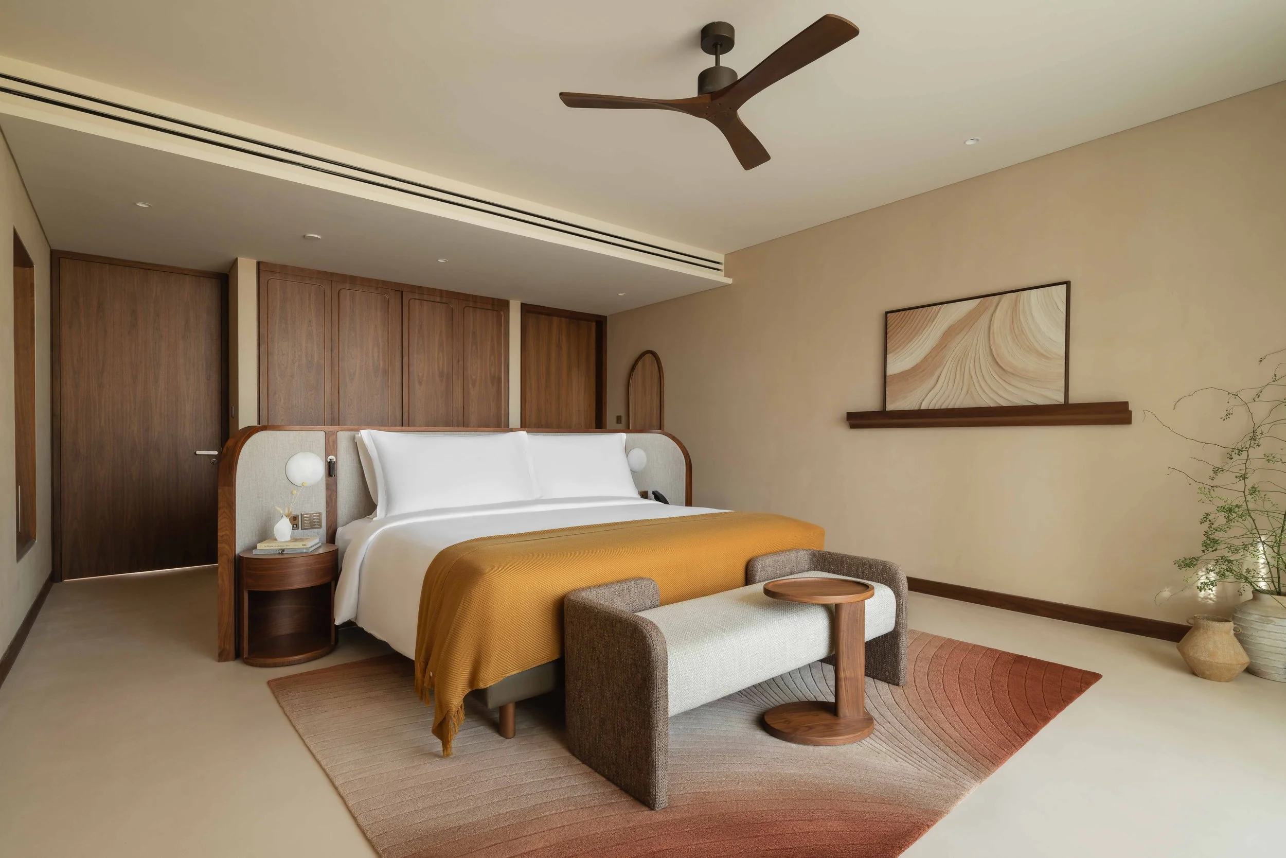 A modern bedroom with a bed, upholstered headboard, and yellow blanket, a small armchair at the foot, a wooden wall panel, and a minimalist art piece on the wall, with a ceiling fan and soft lighting.