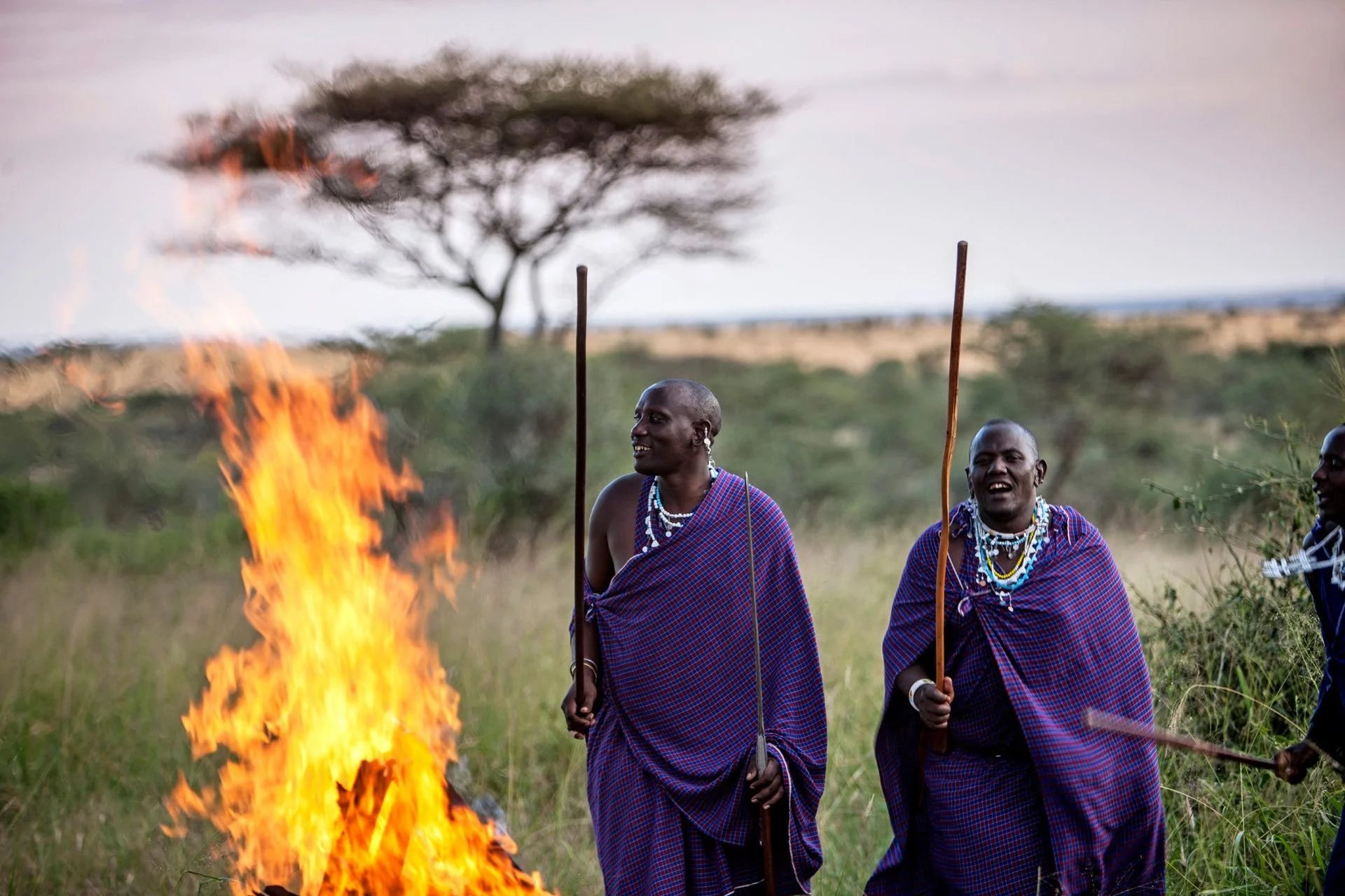 Two Maasai men wearing traditional shukas and beaded jewelry standing near a large fire in an open grassy landscape, with a tree in the background.
