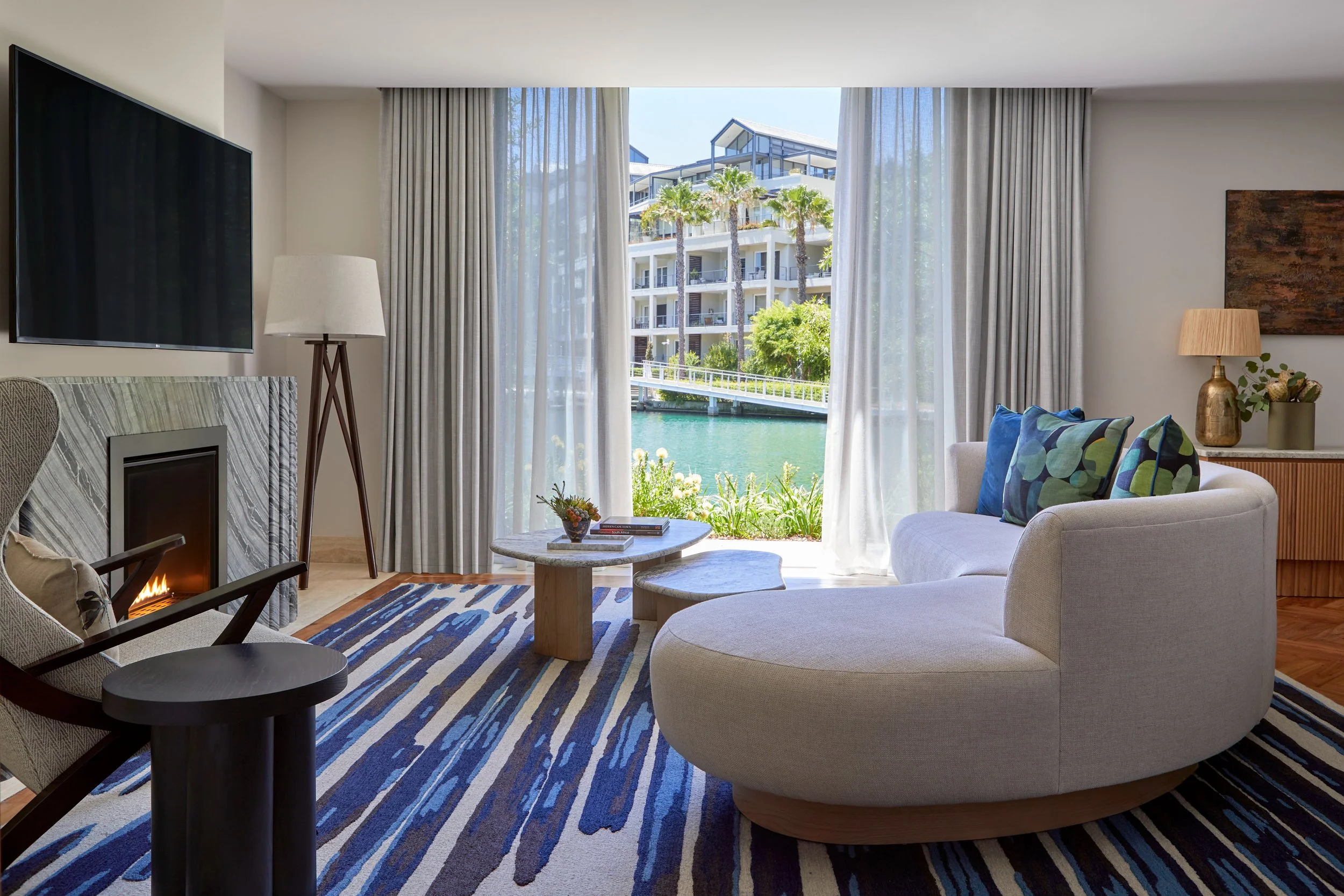 Living room with a large window showing an outdoor view of a waterway, palm trees, and modern buildings. The room has a white curved sofa with patterned pillows, a marble coffee table with books and a small plant, a patterned blue and white rug, a fireplace, and various lamps and artwork on the walls.