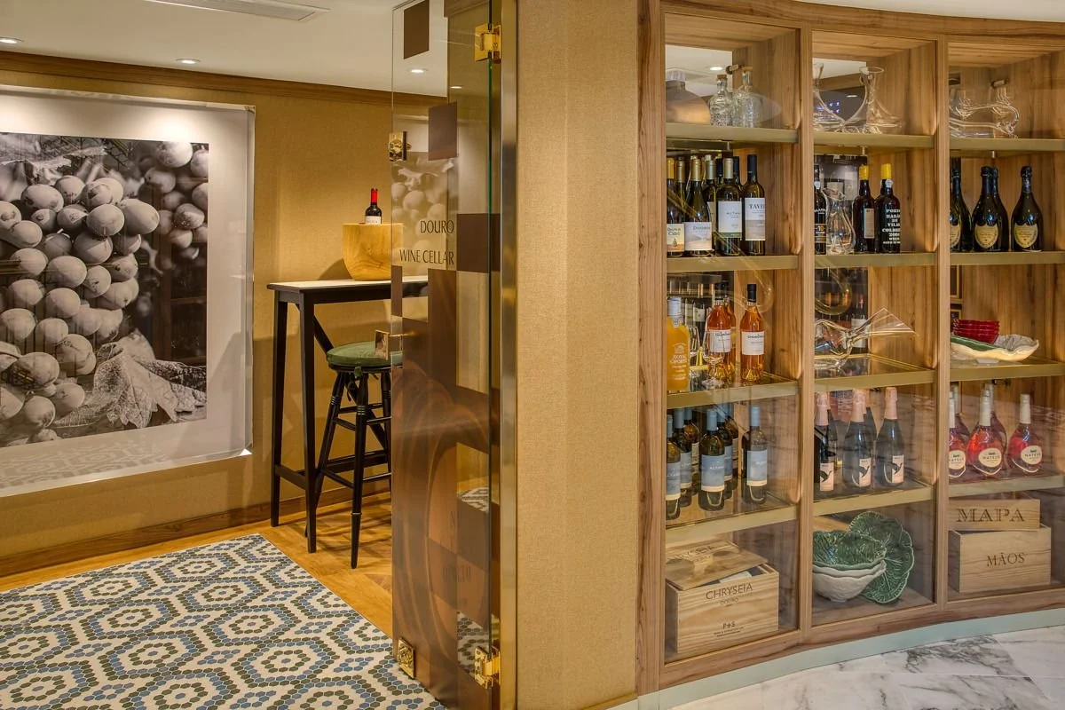 View of a wine cellar corner with glass-fronted wooden shelves filled with bottles of wine and spirits, a large black-and-white food photograph on the wall, a small table with a bottle of wine and a bowl, and a patterned rug on the wooden floor.