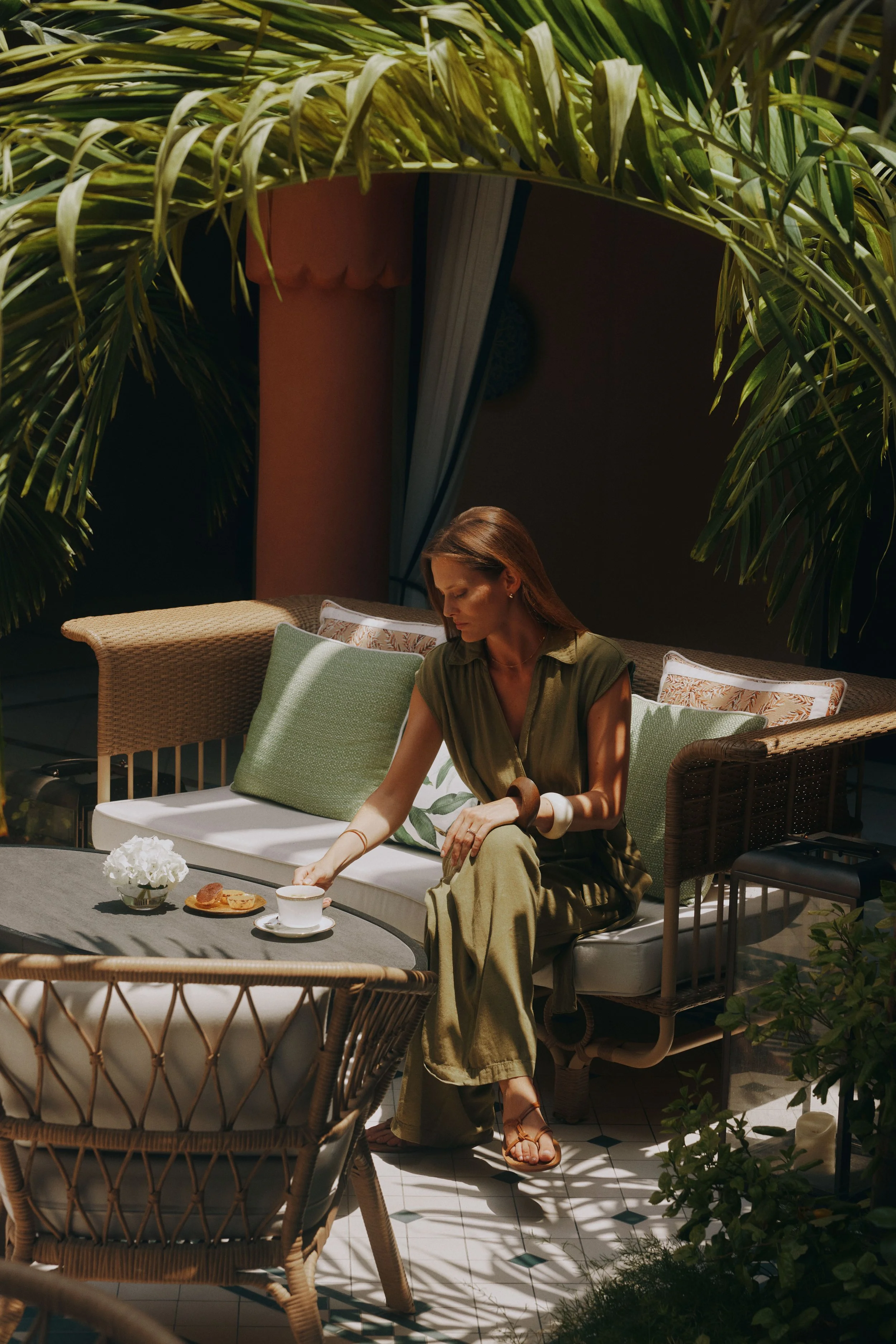 A woman sitting on a wicker outdoor sofa with green pillows, reaching for a cup of coffee or tea on a gray outdoor table. The table has a small white bowl of white flowers and a plate with some baked goods. The area is shaded by large green palm leaves, with sunlight creating patterns on the floor and furniture.