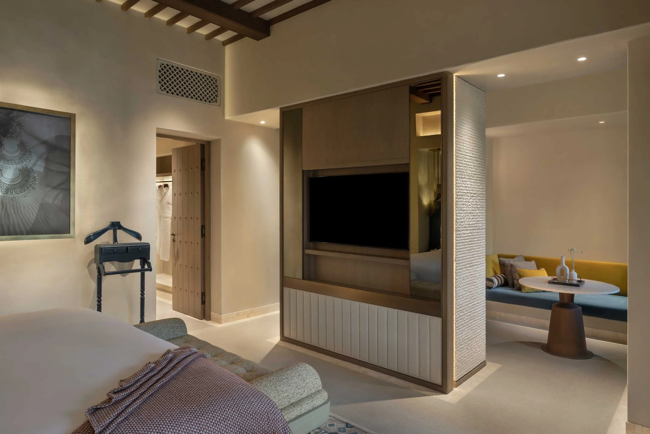A modern hotel suite with a flat-screen TV on a wooden partition, a seating area with a yellow couch and a round table, decorative pillows, and a partial view of a bedroom with a bed and checked blanket.