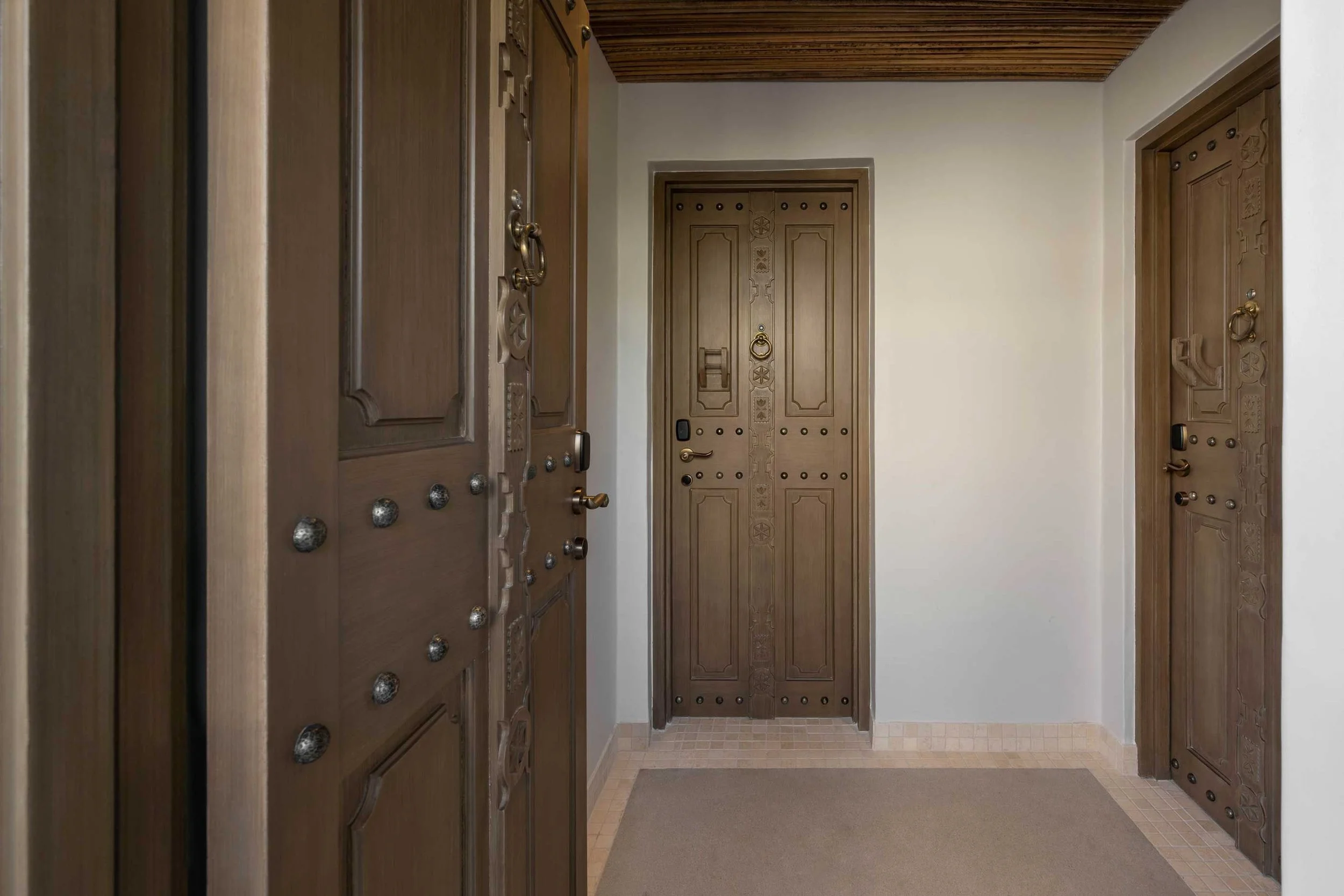 Hallway with wooden doors on both sides, some doors decorated with intricate carvings and metal studs, and a ceiling with wooden paneling.