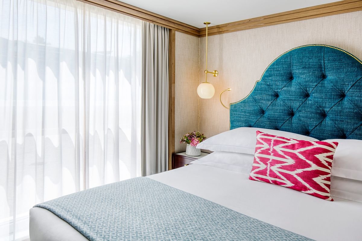 A hotel bedroom with a large blue upholstered headboard, white bedding, a red and white patterned pillow, a side table with a vase of flowers, and a hanging gold and white light fixture, with sheer curtains covering a window.
