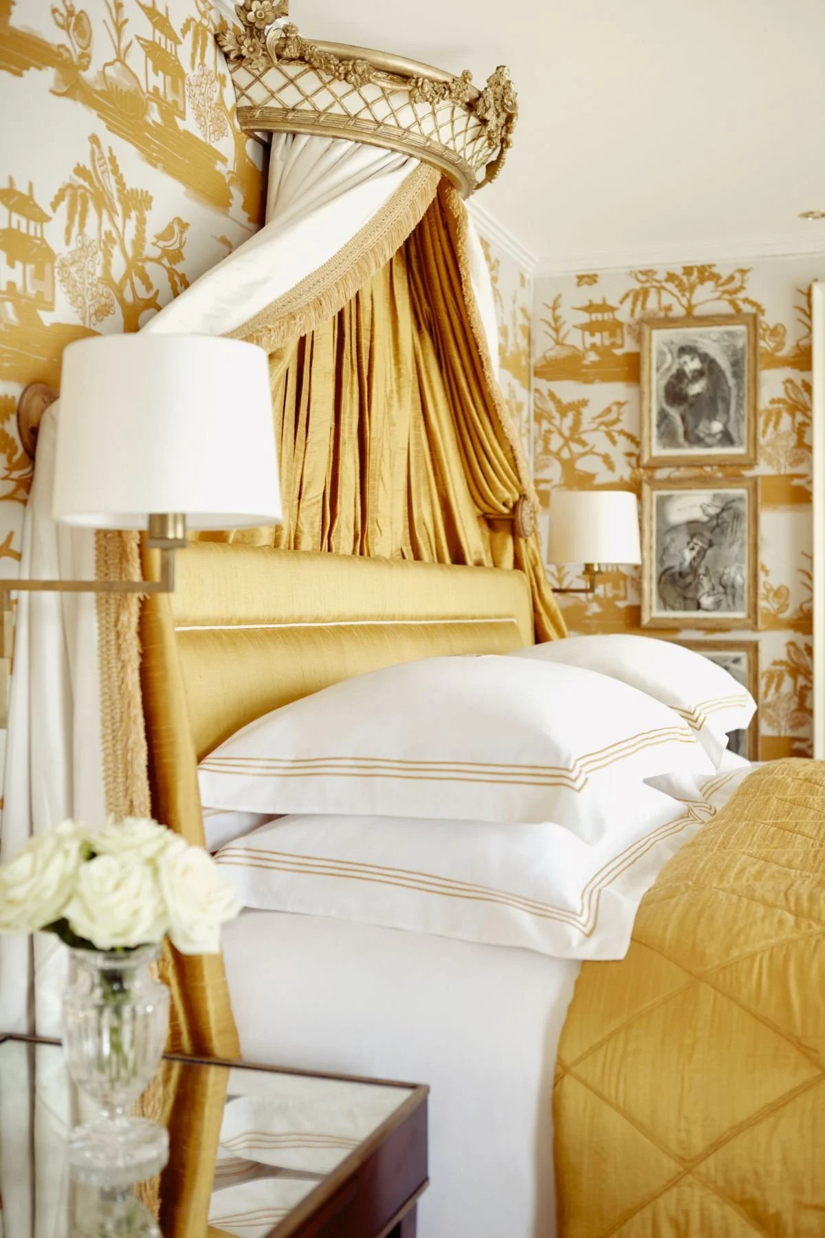 A luxurious bedroom with a gold and white color scheme featuring a canopy bed with ornate gold drapes, a gold upholstered headboard, white pillows with gold accents, and gold wallpaper with Asian-inspired motifs. There are matching lamps and framed artwork on the wall.