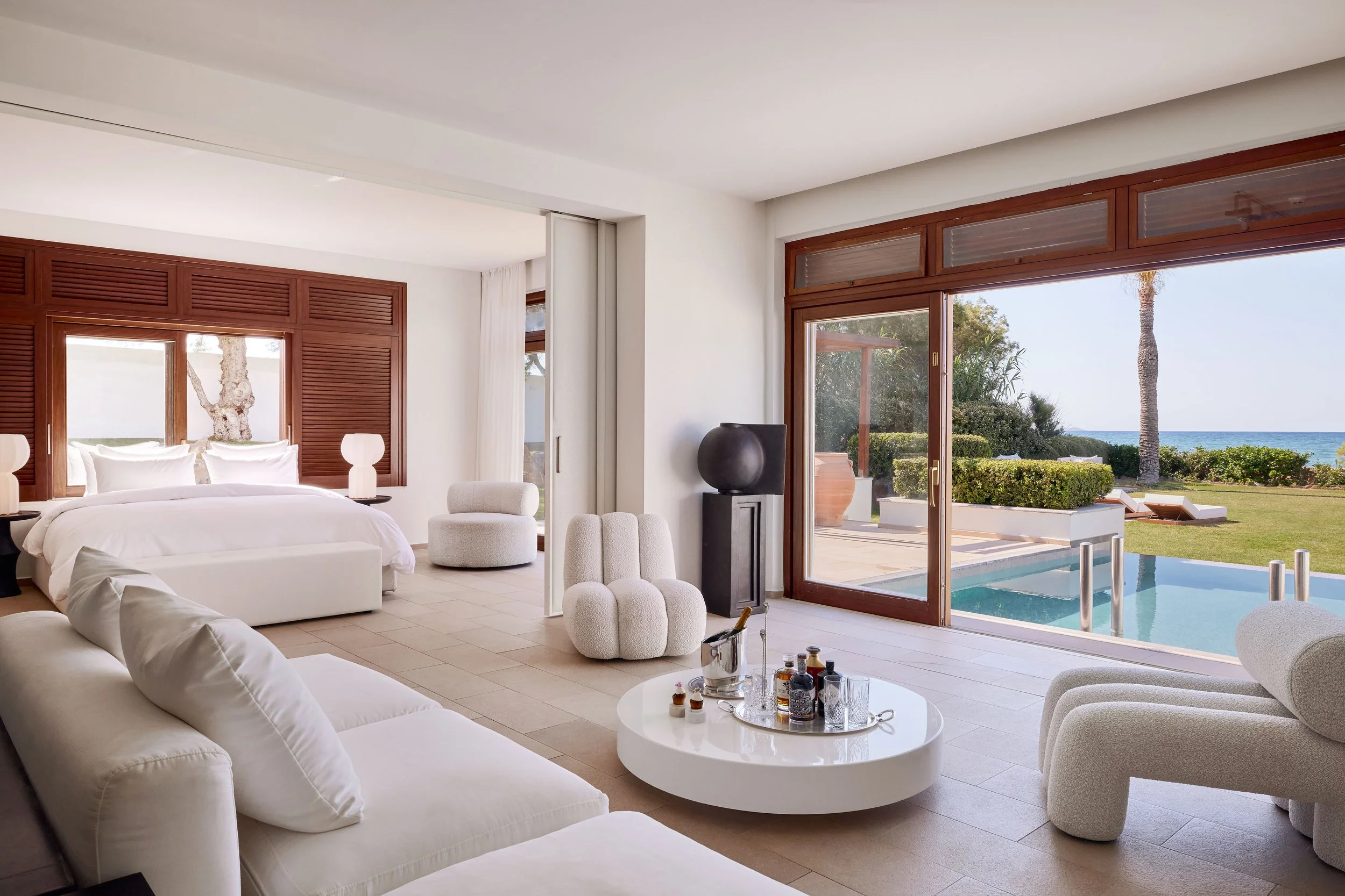 Modern living room with large glass windows and doors overlooking a yard with palm and other trees, and the ocean in the background. The room has white furniture, including a sofa and curved armchairs, and decorative items like lamps and a coffee table with bottles.
