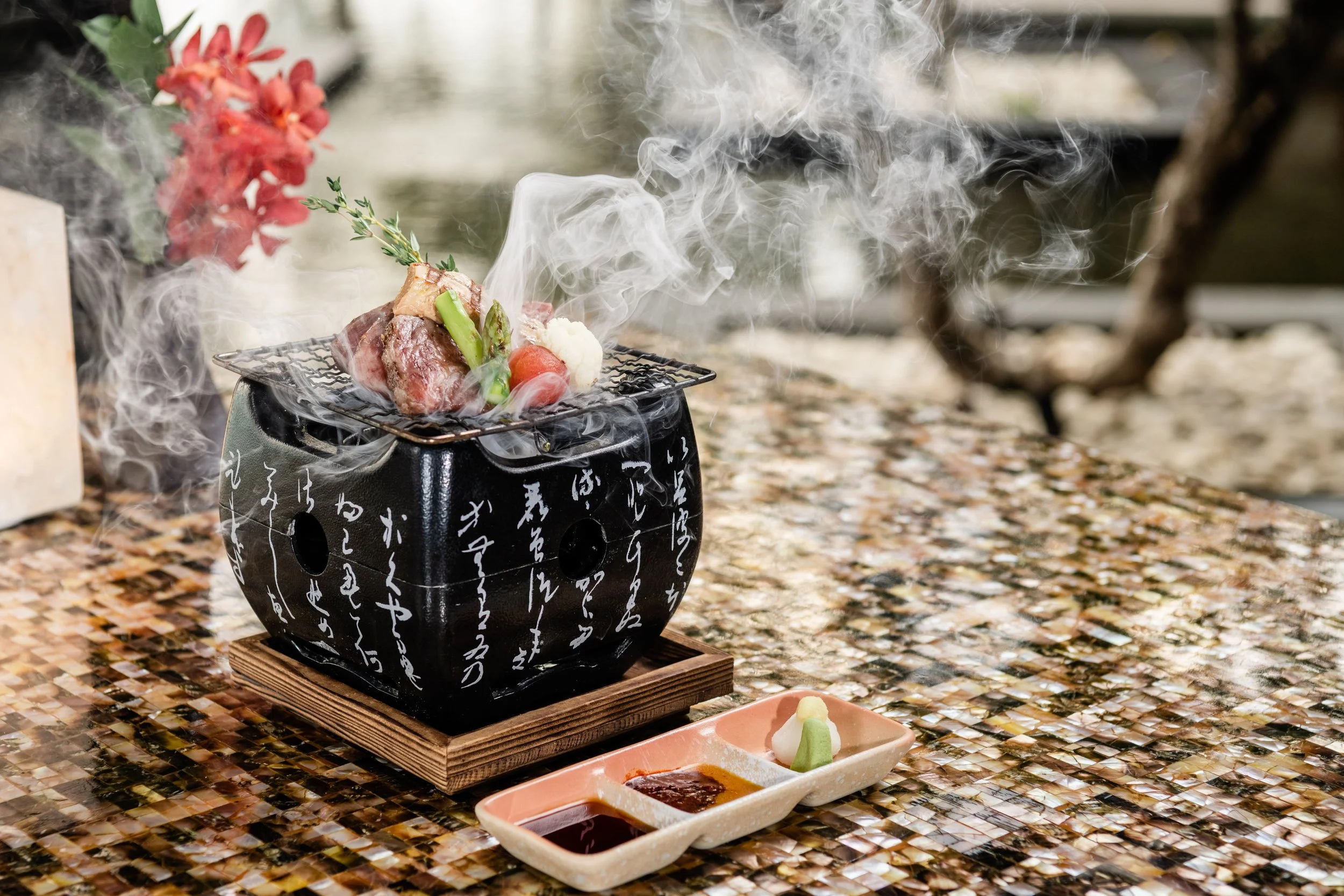 Hot stone grill with raw meat, vegetables, and herbs, emitting smoke, placed on a wooden stand, with dipping sauces and wasabi on the side.