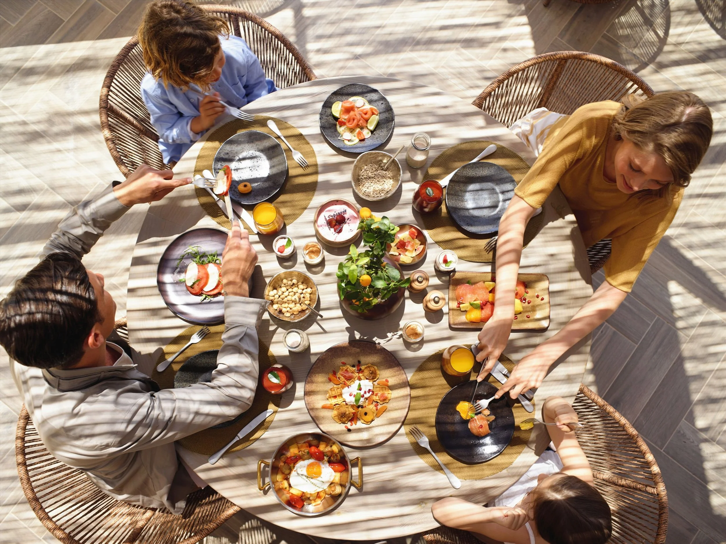 A top view of a breakfast scene with several people gathered around a table. The table is filled with various breakfast dishes including toast, eggs, fruit, oatmeal, and beverages like orange juice and coffee. The setting appears to be indoors with natural sunlight casting shadows.