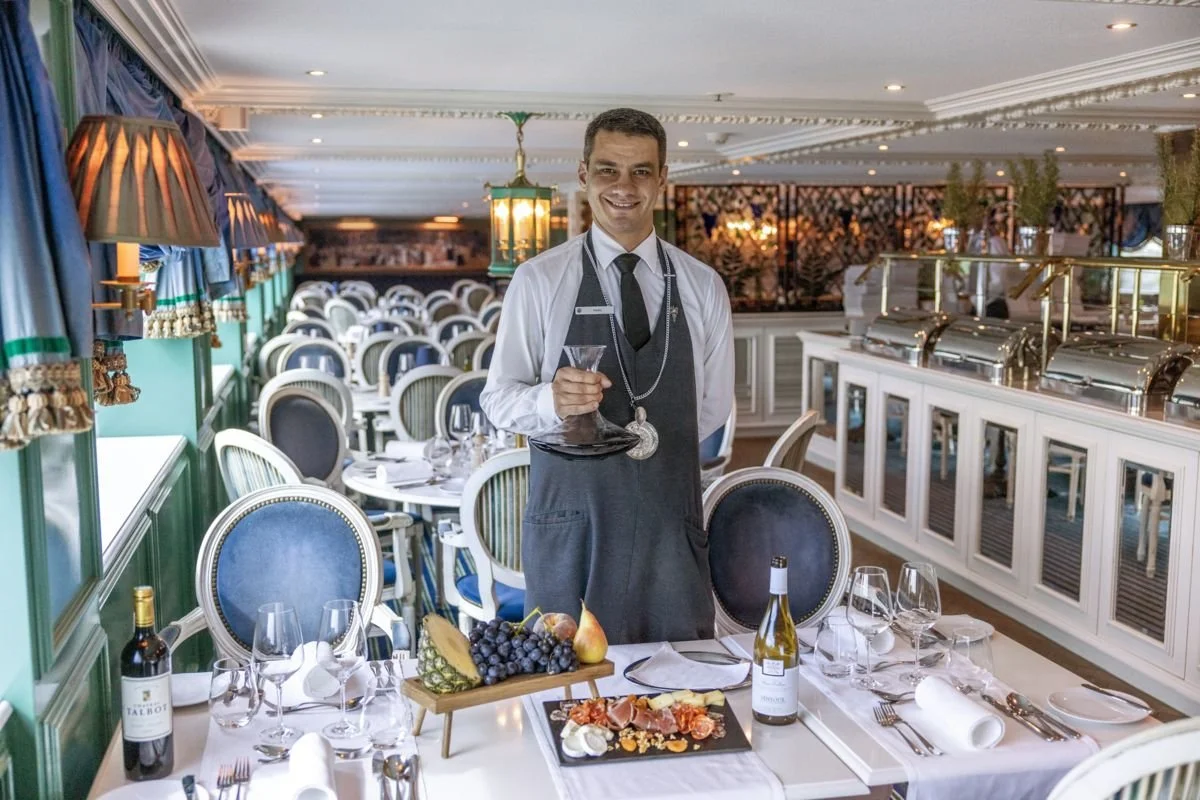 A smiling waiter in a formal uniform holding a martini glass at a dining table in a decorated restaurant with neatly arranged chairs, wine bottles, and a platter of grapes and cheese.