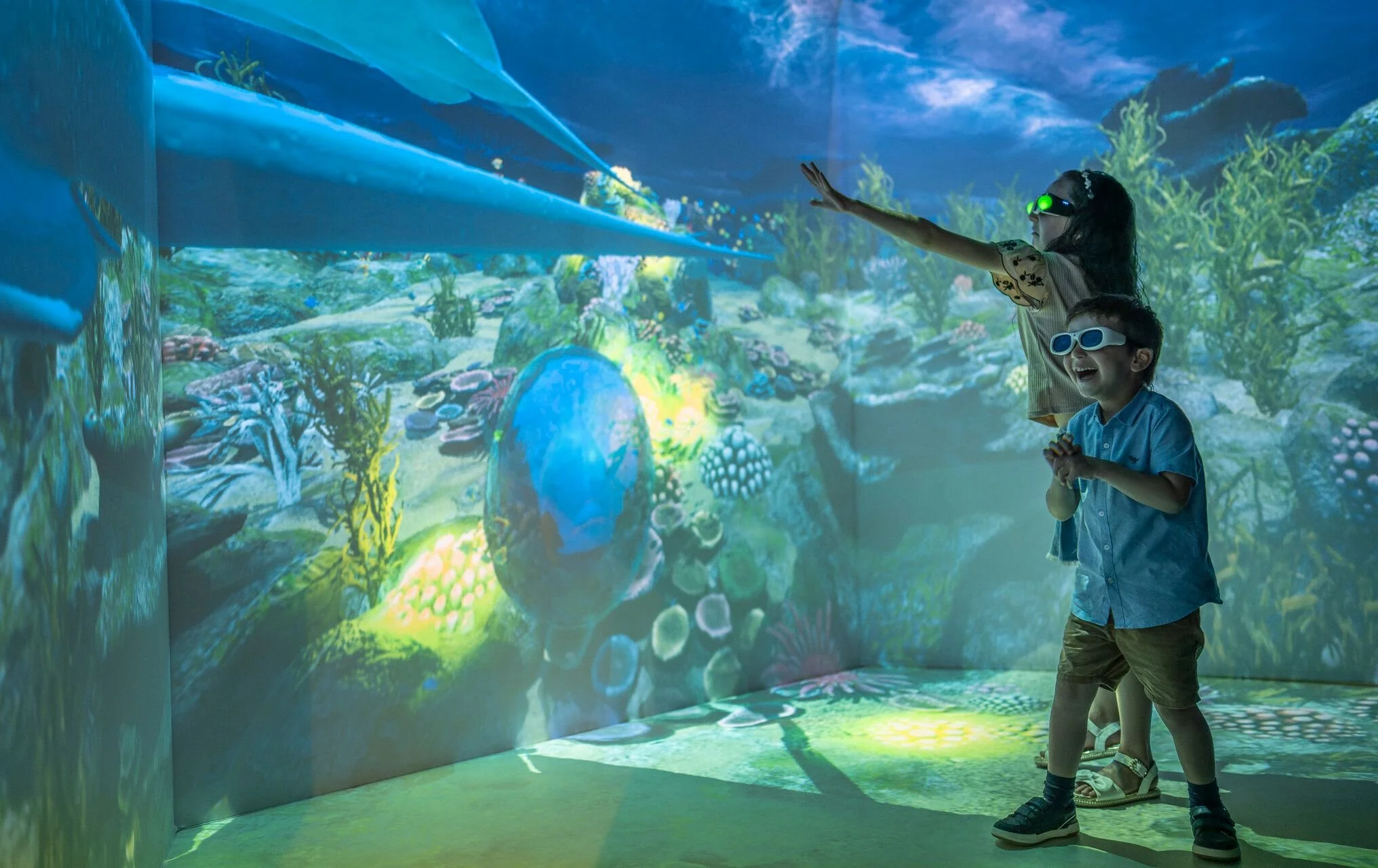 Two children wearing 3D glasses enjoy an immersive digital underwater scene with coral reef, fish, and water effects at an aquarium or digital exhibit.