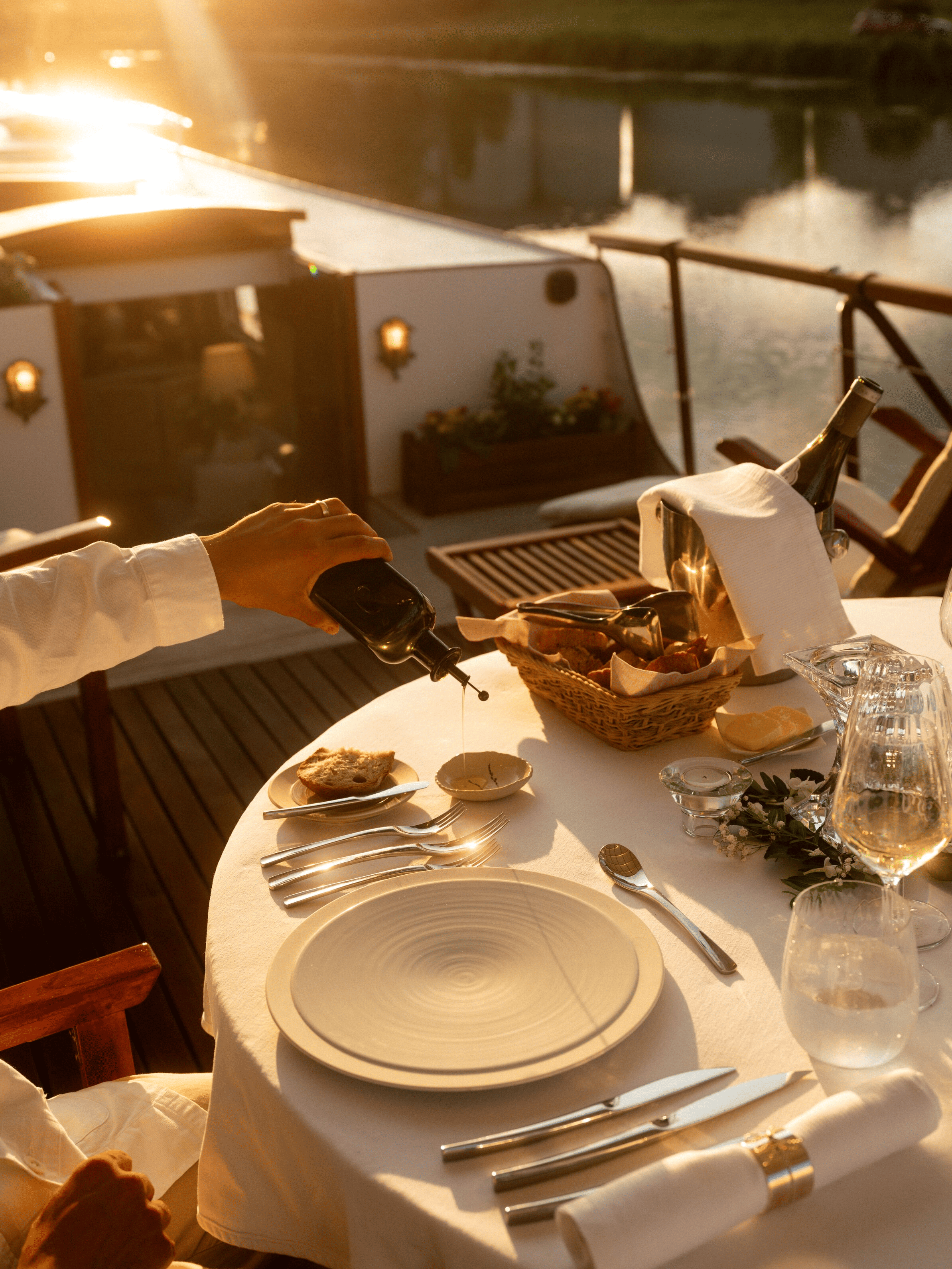 A table set for dinner on a boat with plates, glasses, silverware, a basket of bread, and a wine bottle in an ice bucket, overlooking water with a dock in the background during sunset.