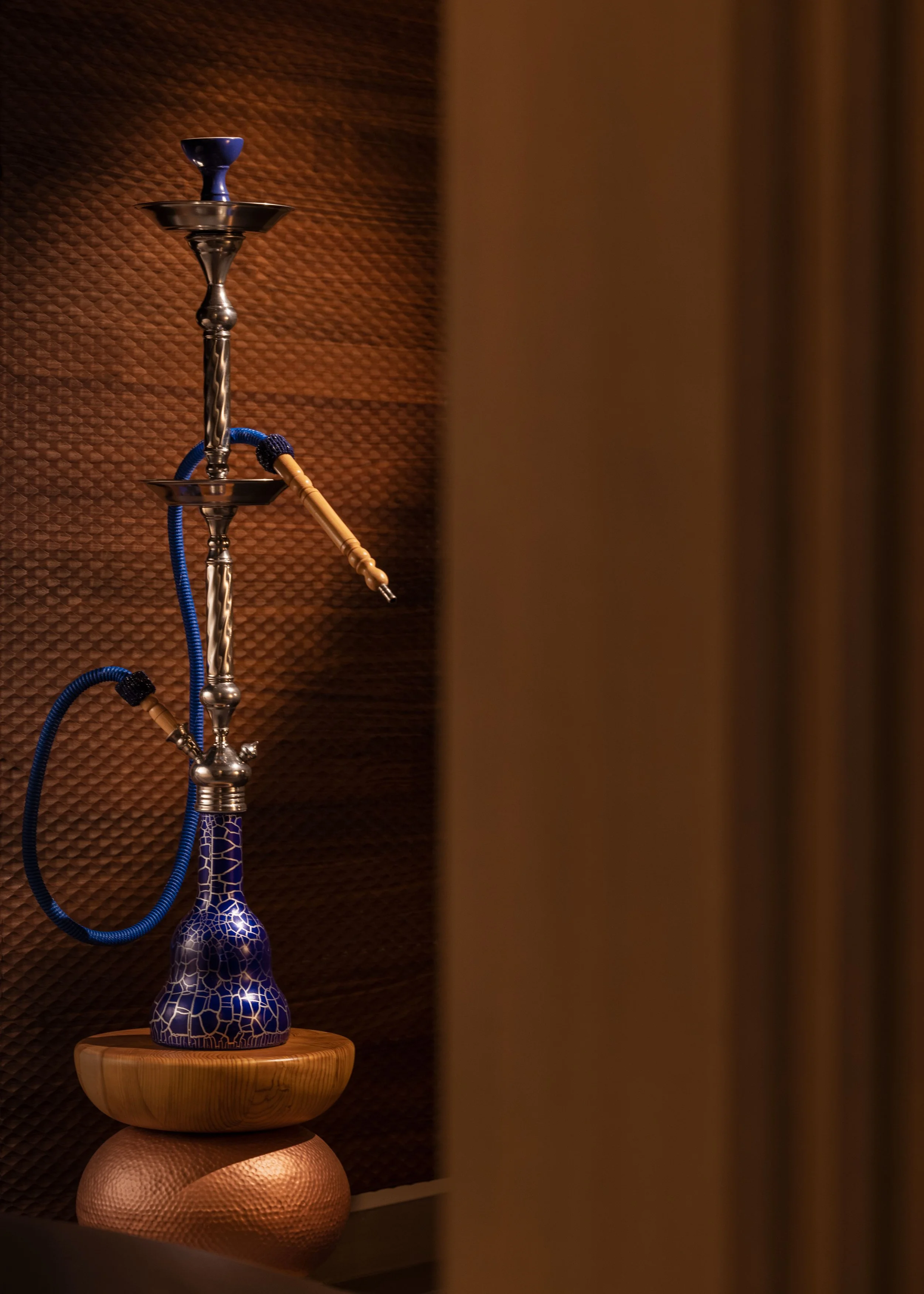 A tall hookah with a blue and white mosaic-patterned base, metal stem, and blue hose, standing on a wooden surface against a textured brown background.