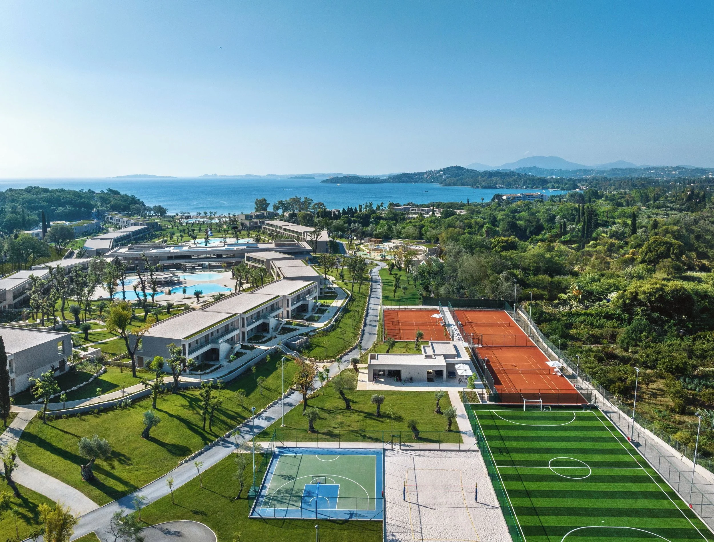 Aerial view of a modern residential community by a large body of water with mountains in the background. The image shows swimming pools, tennis courts, green lawns, and buildings among trees.
