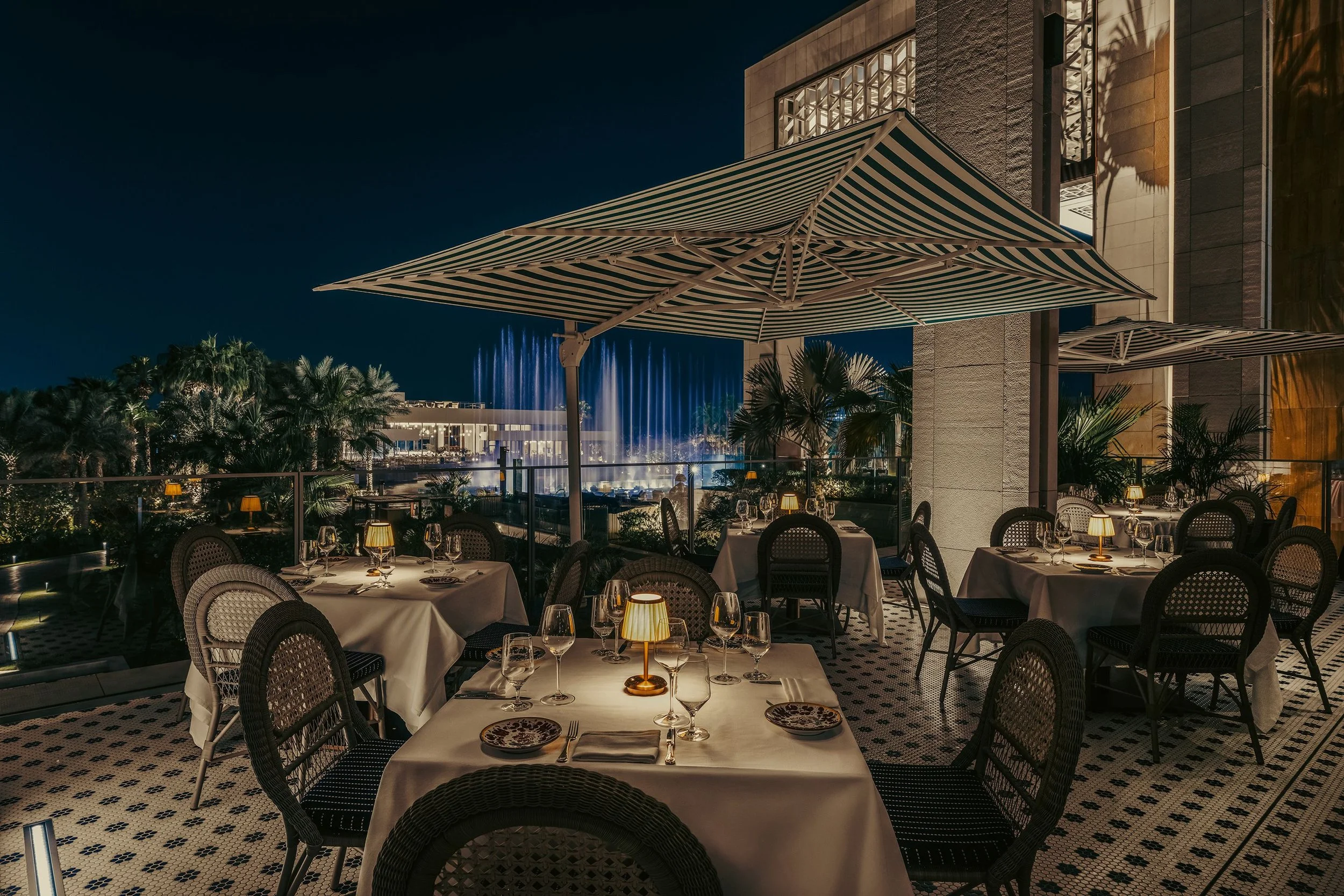 Outdoor restaurant dining area at night with tables set for a meal, large striped umbrellas, palm trees, and a fountain in the background.
