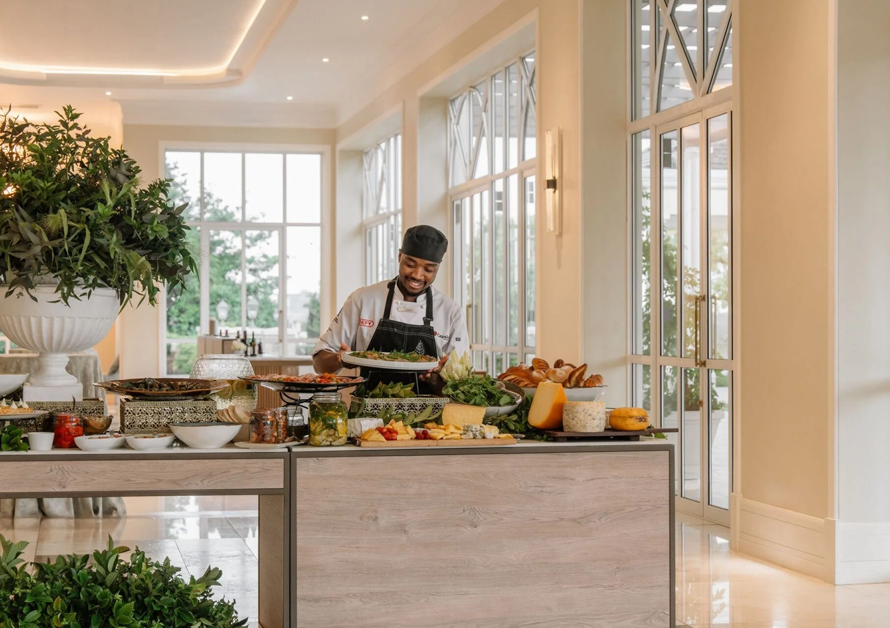 A chef smiling while preparing food at a buffet table with cheeses, bread, and appetizers in a bright indoor setting with large windows and greenery outside.