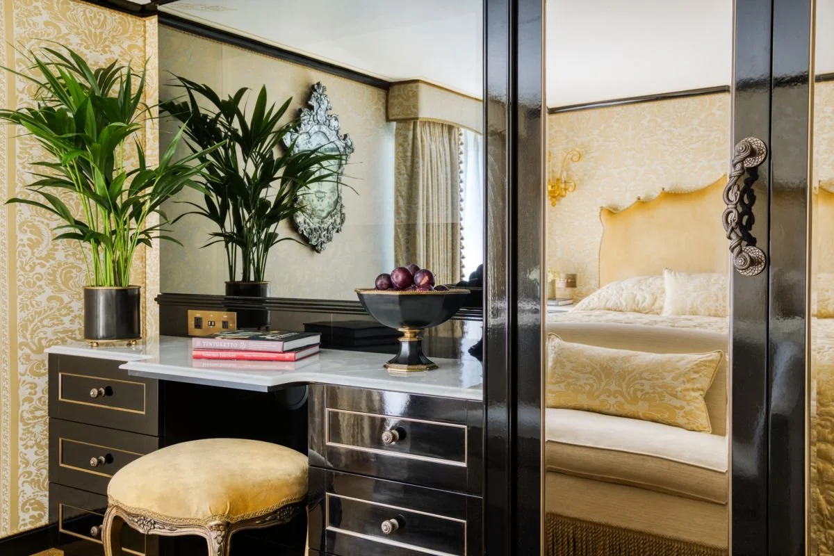 A vanity area in a luxurious bedroom with gold and black decor, featuring plants, books, a bowl of red grapes, a mirror, and a cream upholstered bench.
