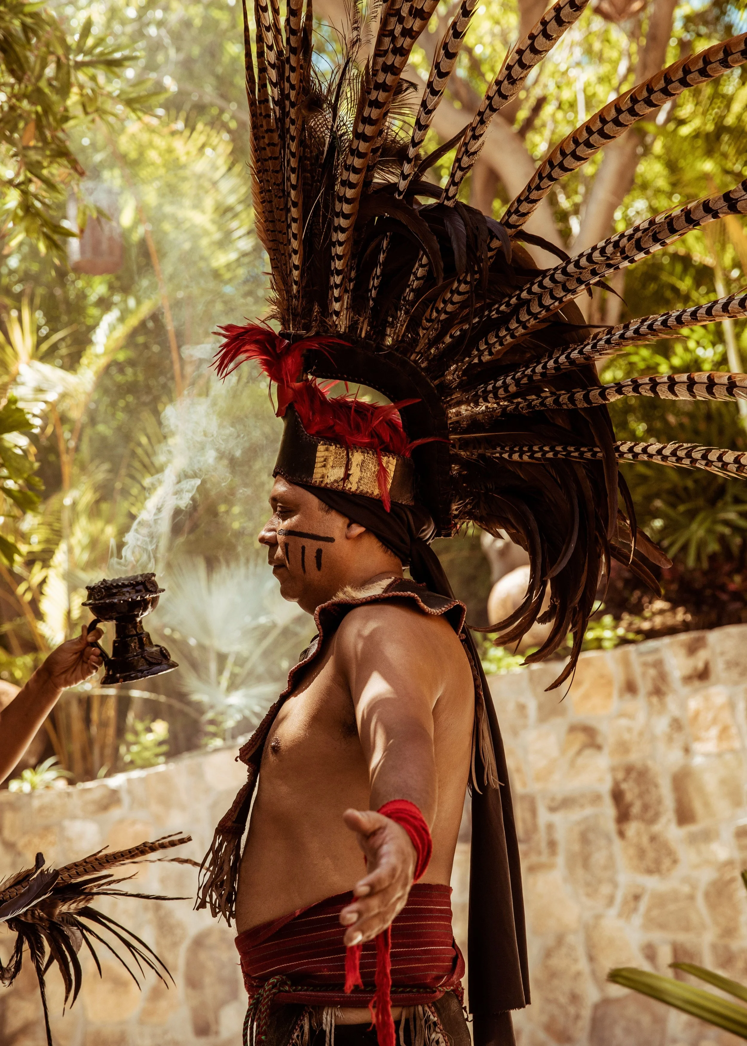 An indigenous man wearing traditional headdress with large feathers, face paint, and red accessories, holding a ceremonial object and performing a ritual outdoors surrounded by trees.