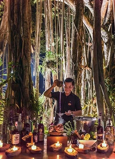 A man preparing drinks at a bar surrounded by tropical plants and candles.
