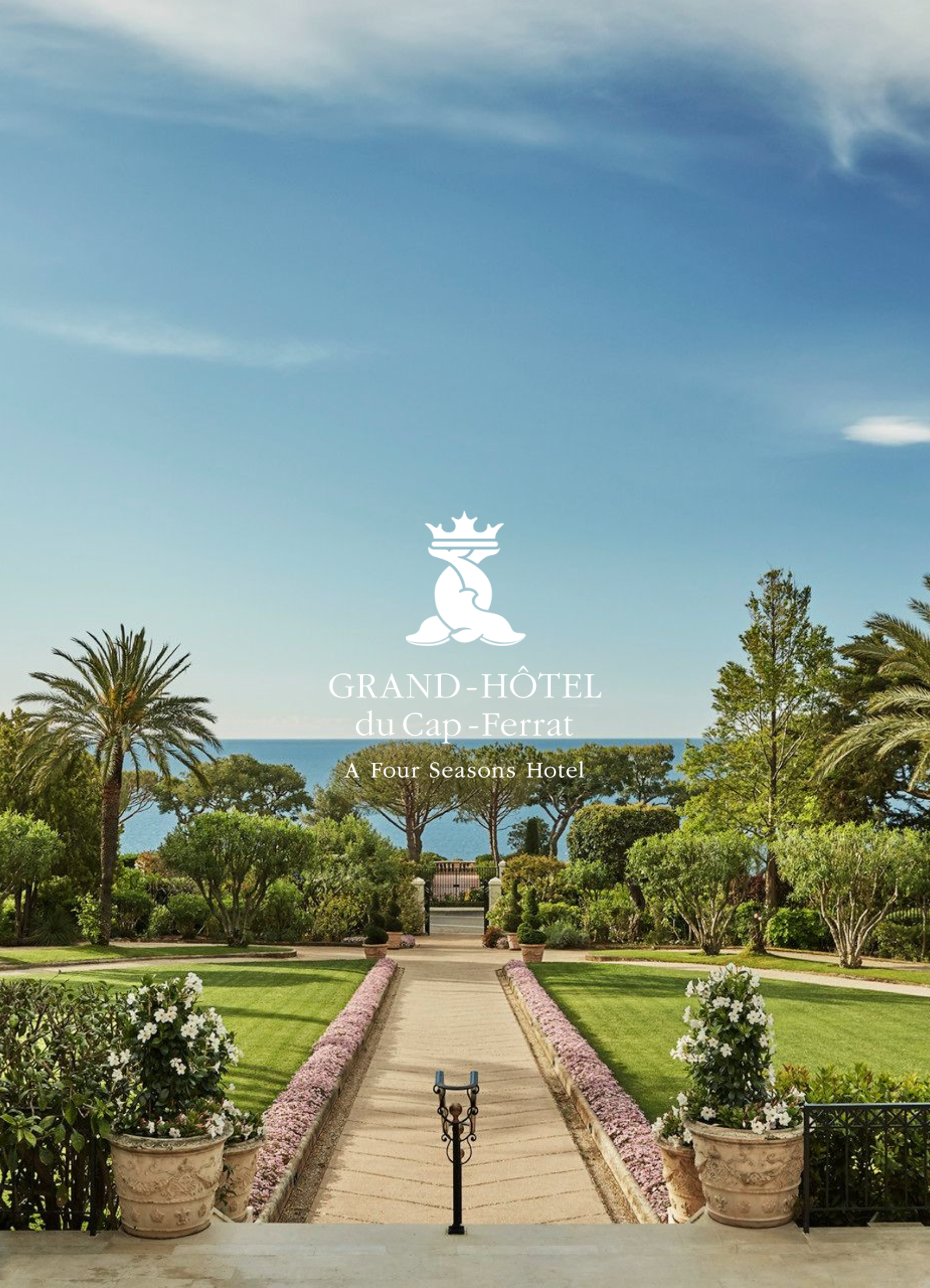 A landscaped garden with a pathway lined with pink flowers leading to lush green trees, shrubs, and a view of the ocean in the background, with the logo and text of Grand-Hôtel du Cap-Ferrat at the top.