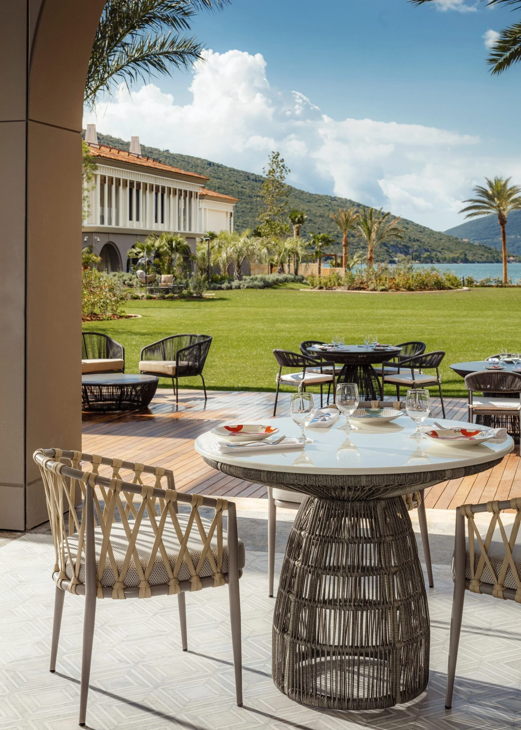 A view of a luxurious outdoor dining area with a round table set with glasses and plates, surrounded by modern chairs with woven designs. Beyond the patio, there is a well-manicured lawn with lush greenery, palm trees, and a scenic view of water and mountains under a blue sky with clouds.