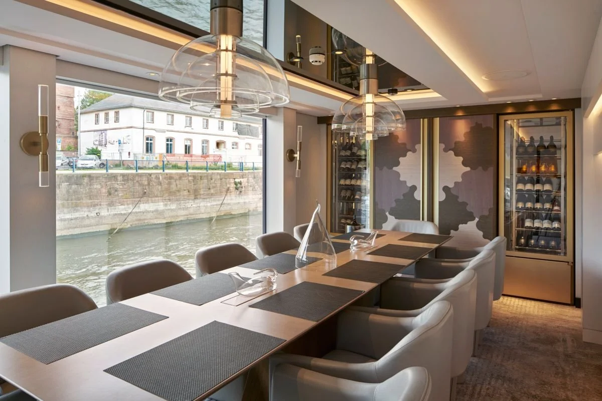 Interior of a modern restaurant with a long dining table, grey chairs, and large glass wine coolers, overlooking a canal through large windows.