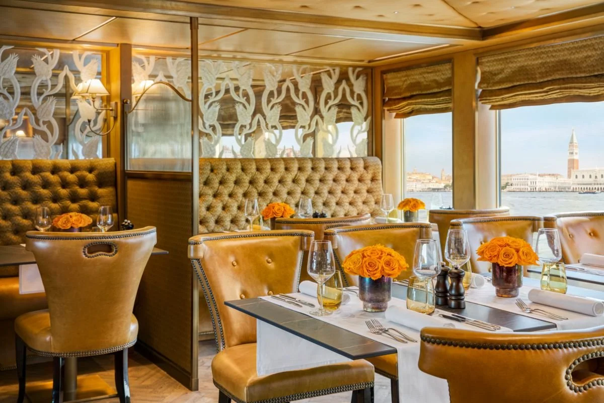 Elegant restaurant interior with a view of a city and water outside large windows, featuring beige tufted seating, wooden accents, and tables decorated with orange roses in vases, wine glasses, silverware, and napkins.