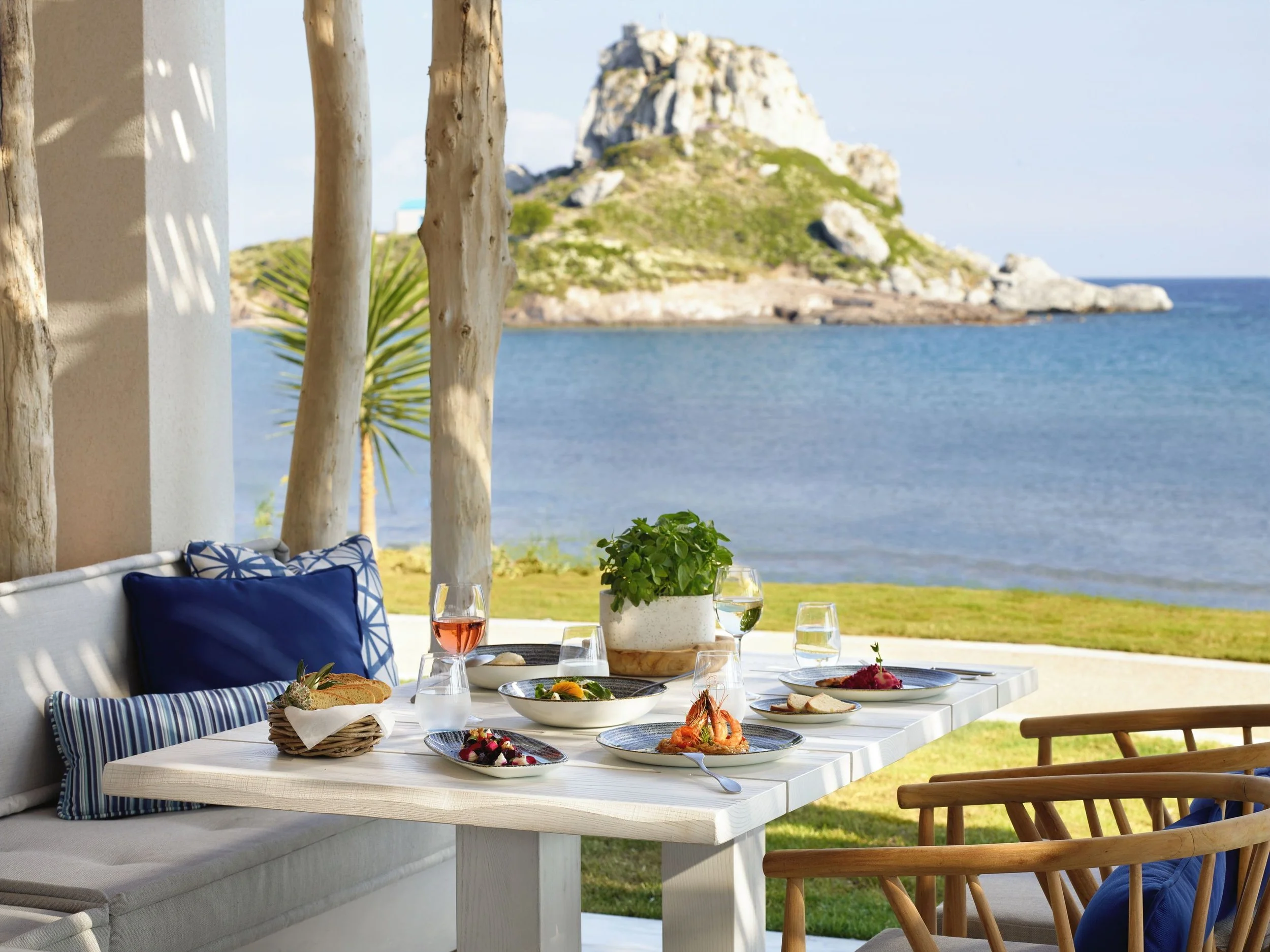 Dining table with food and drinks on a patio overlooking a lake and a rocky hill.