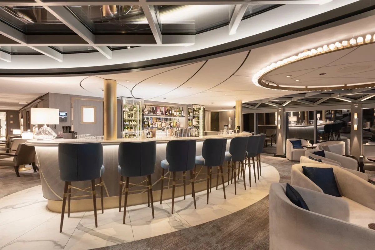 Elegant hotel lounge with a curved bar, high-backed blue chairs, marble flooring, plush beige sofas with dark cushions, and modern lighting fixtures.