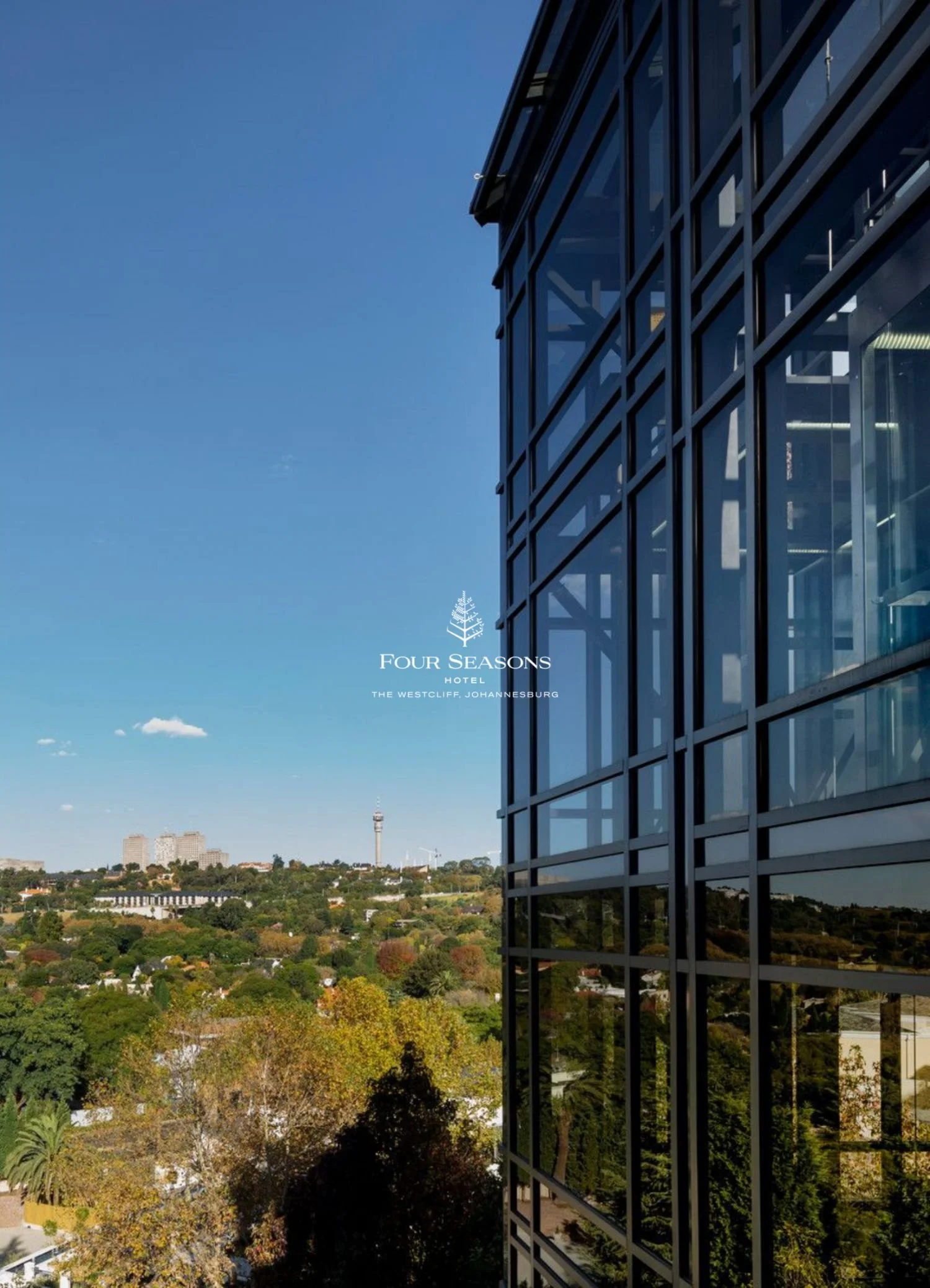 View of a cityscape with a modern glass building on the right side, trees in the foreground, and a clear blue sky in the background.