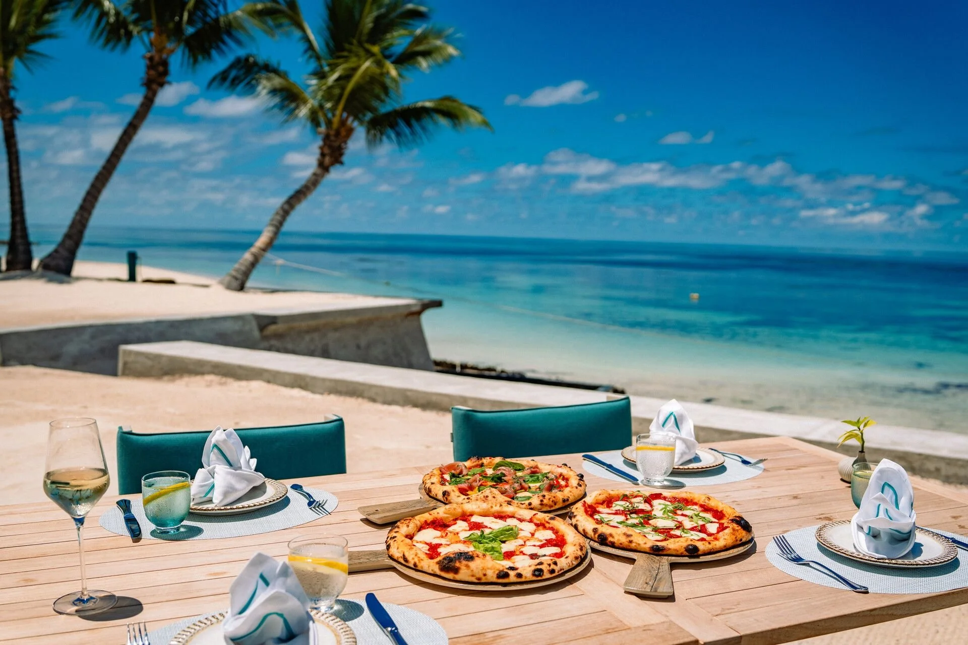 Outdoor dining table with three pizzas, glasses of wine and water with lemon, and silverware set on white napkins, overlooking a beach with palm trees and blue ocean under a partly cloudy sky.