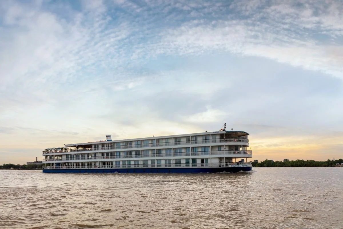 A large river cruise boat sailing on a calm river during sunset, with a partly cloudy sky and distant trees in the background.