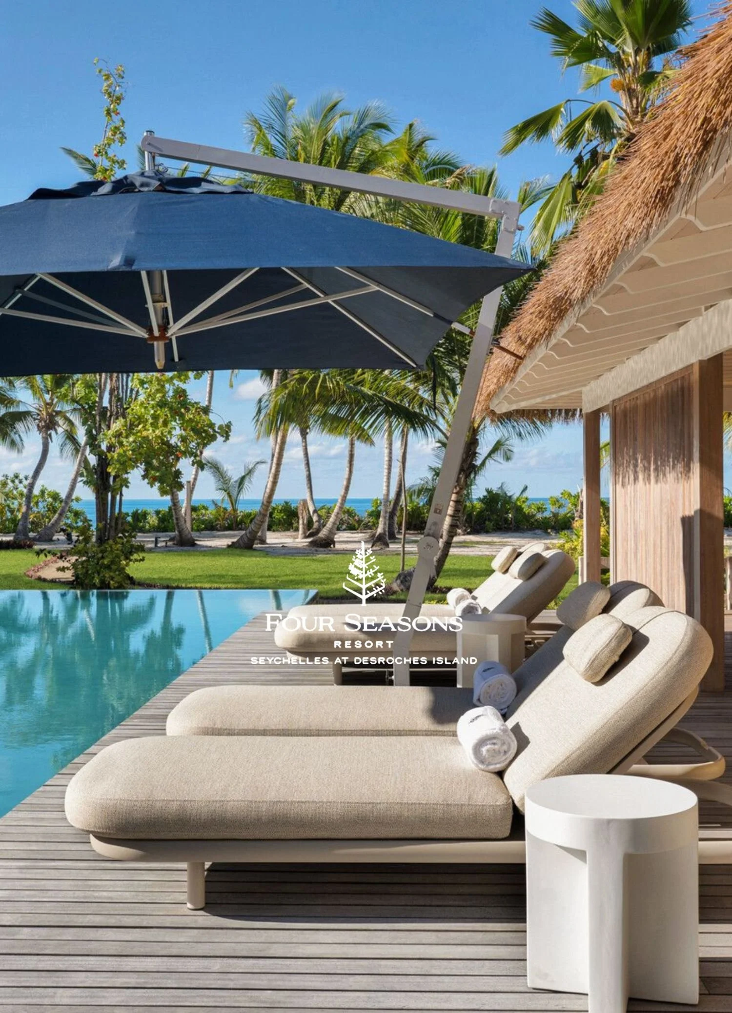 Poolside lounge chairs with rolled towels, white side table, thatched roof structure, large blue umbrella, wooden deck, tropical trees, and a view of the ocean in the background at the Four Seasons Resort in Seychelles at Desroches Island.