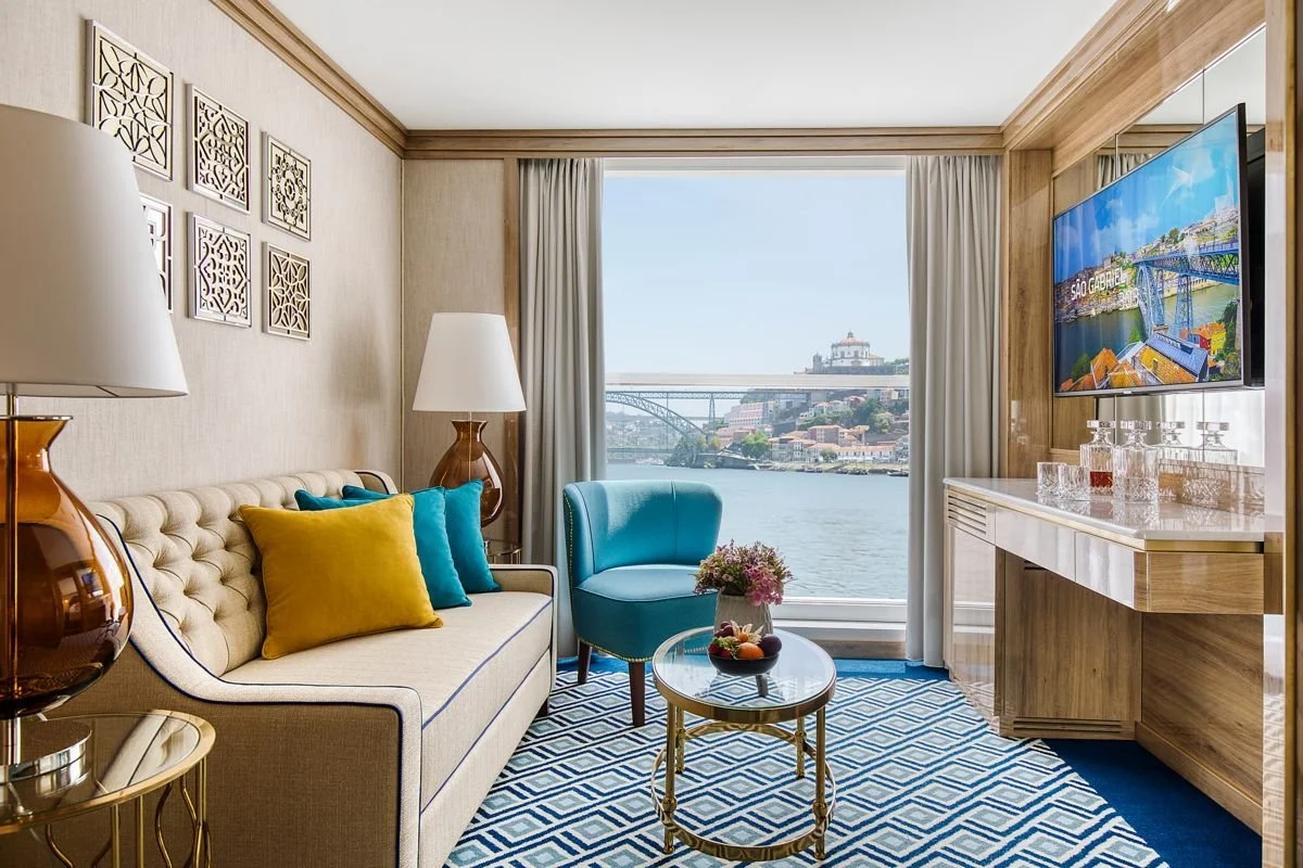 Interior of a hotel room with a sofa, colorful cushions, a blue armchair, coffee table with flowers, a TV on the wall, and a large window overlooking a river, bridge, and cityscape.
