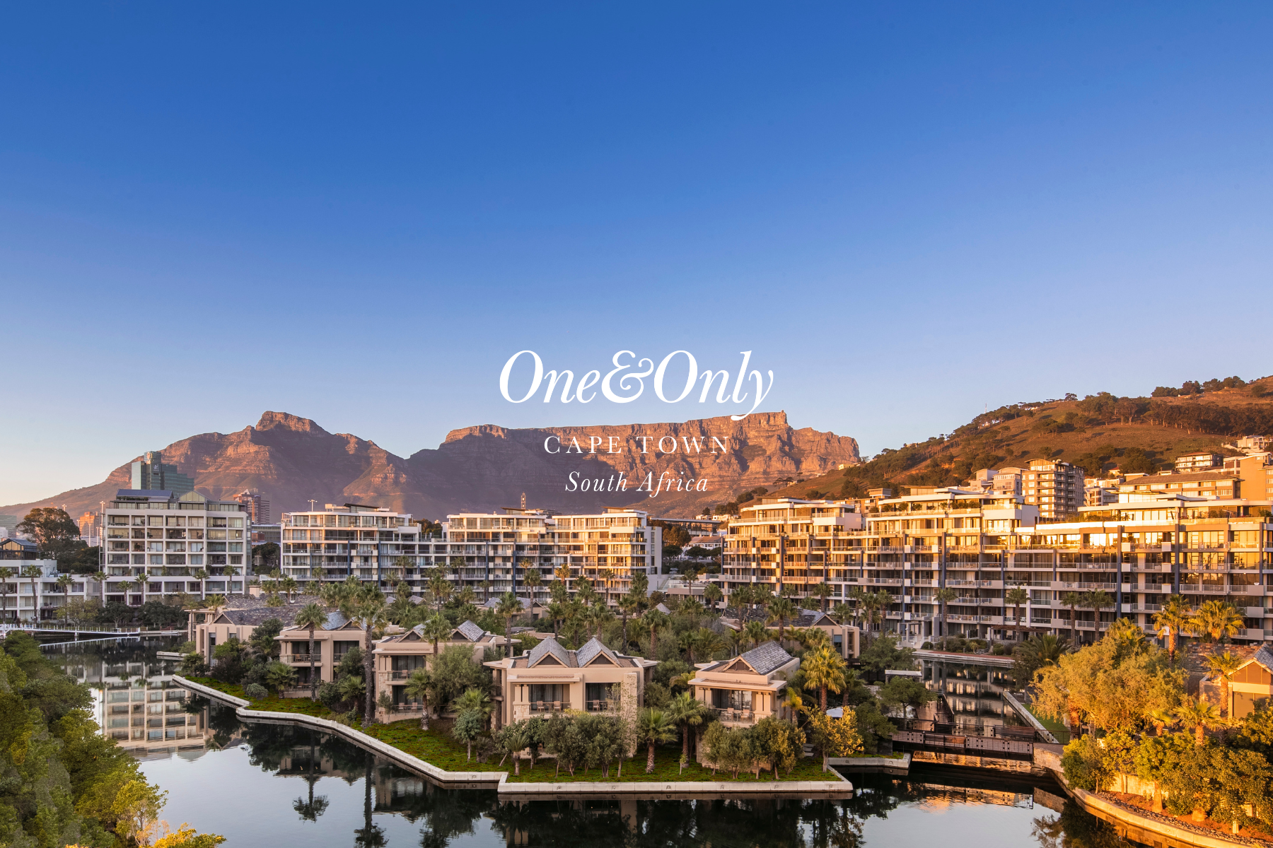 Residential buildings with a body of water in the foreground, surrounded by trees and palm trees, with a mountain range in the background under a clear sky.